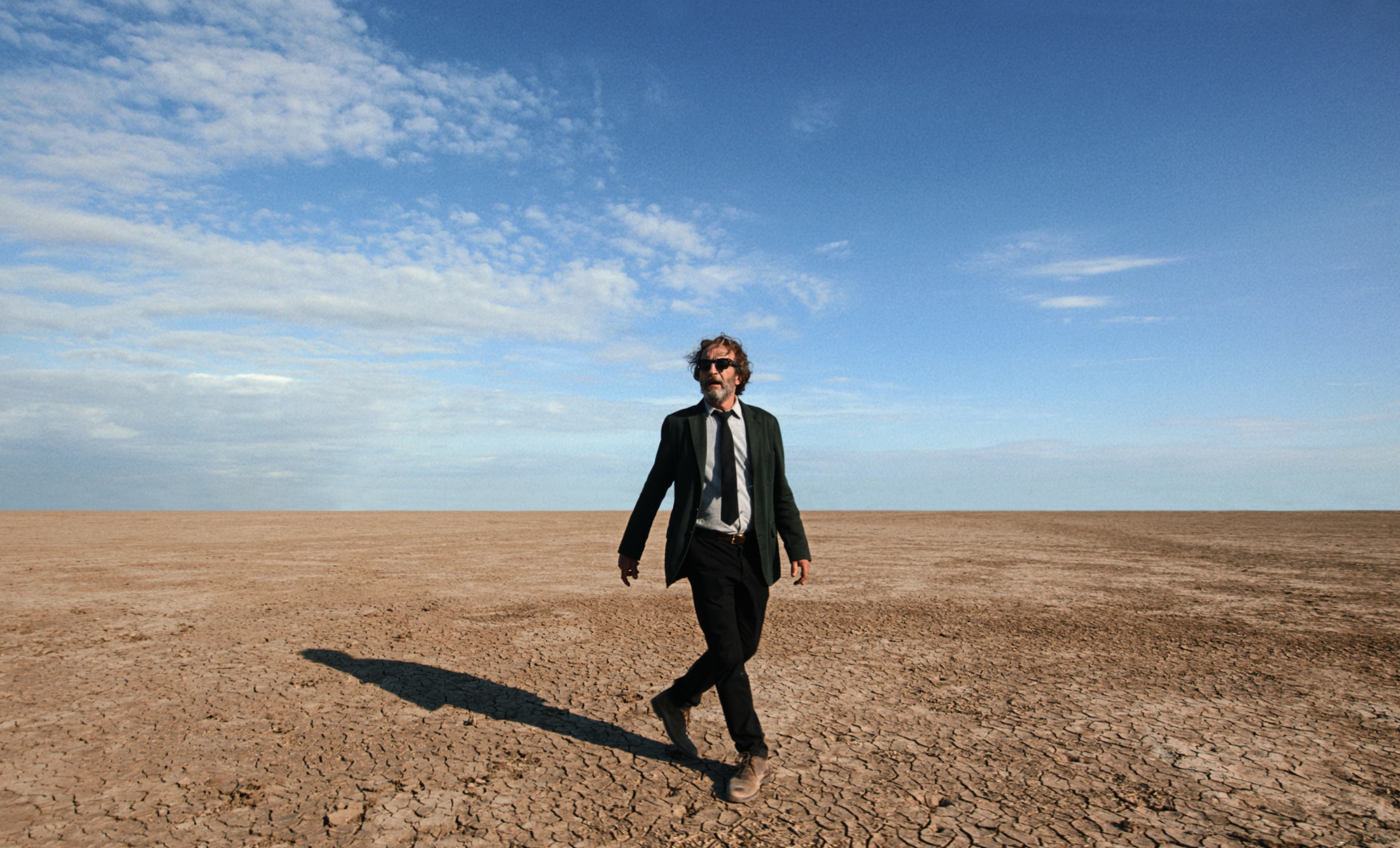 'Bardo' Daniel Giménez Cacho as Silverio Gacho (1) walking in the desert wearing a suit, tie, and sunglasses. His long shadow on the ground behind him.