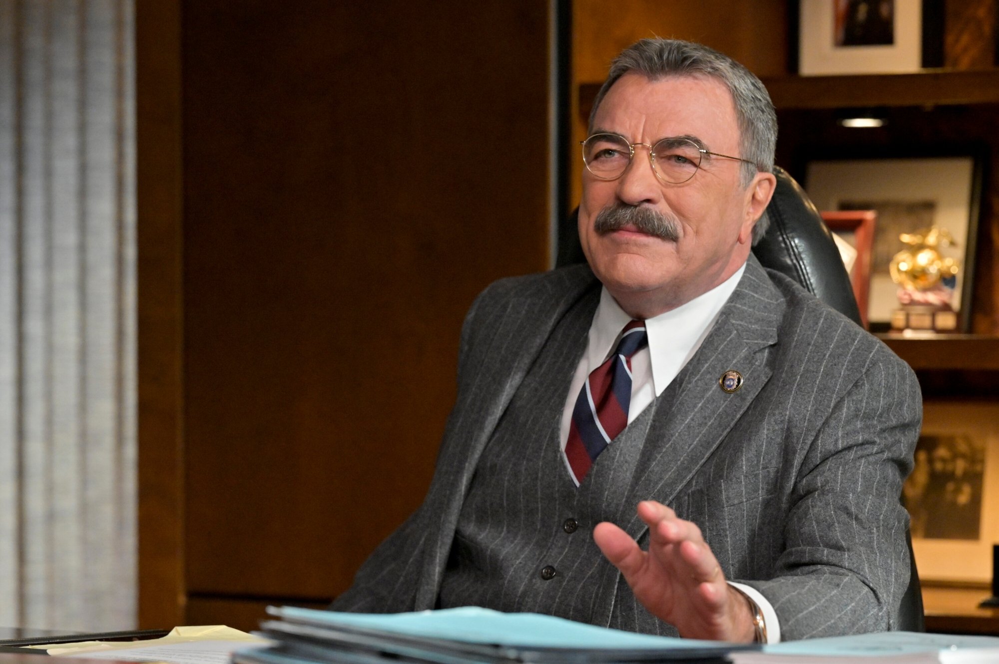 'Blue Bloods' Tom Selleck as Frank Reagan wearing a suit, tie, and glasses. He's sitting at a desk with his hand slightly raised off the table.