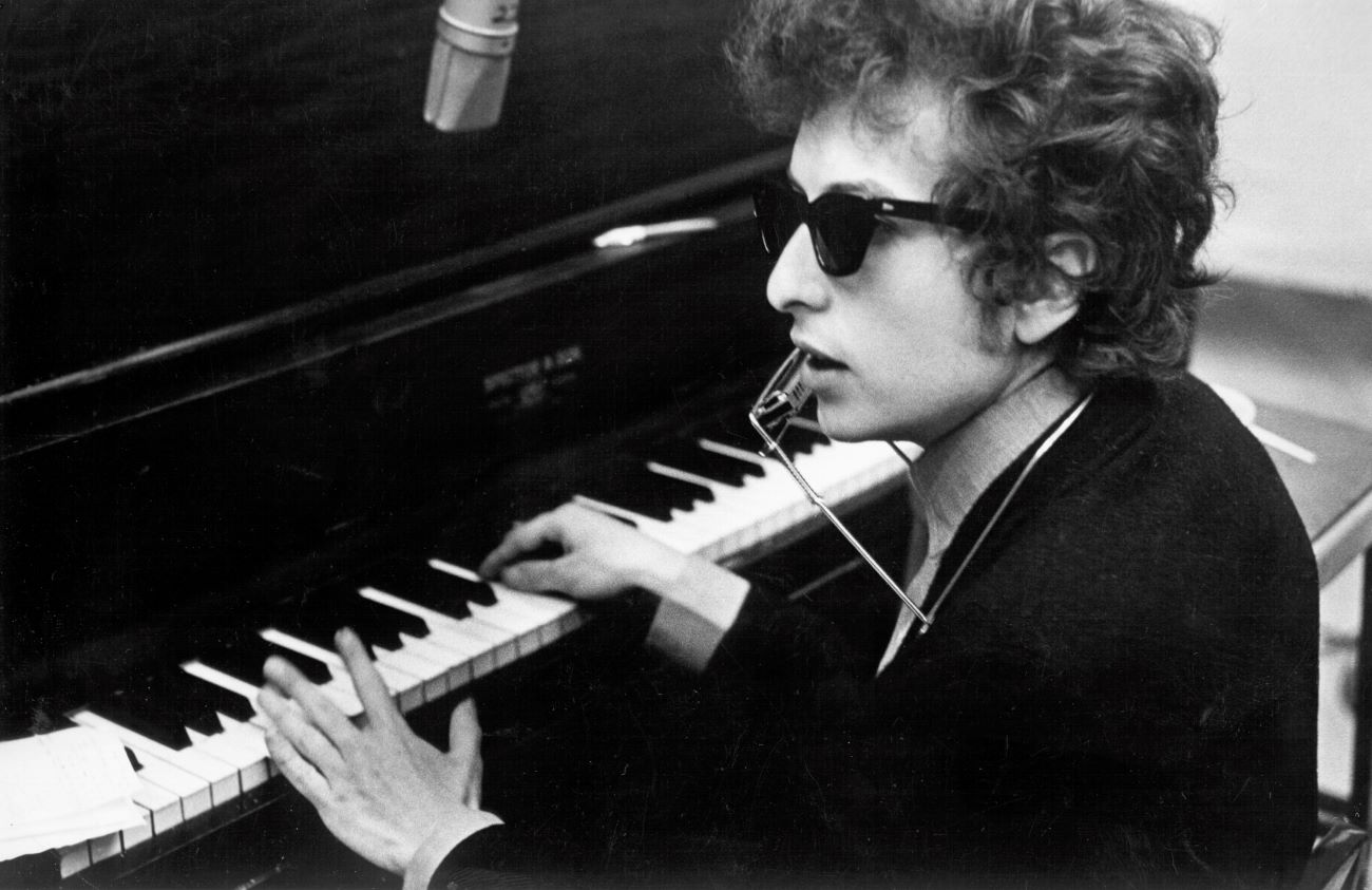 A black and white picture of Bob Dylan wearing sunglasses and sitting at a piano.