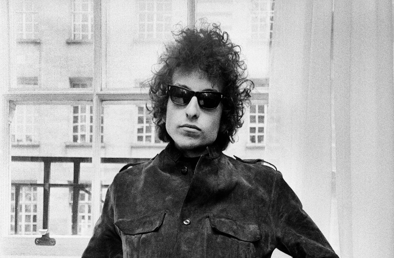 Bob Dylan wears sunglasses and stands in front of a window.