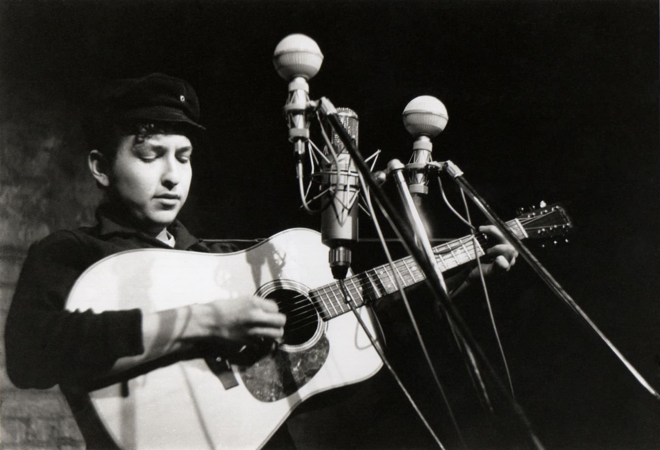 A black and white picture of Bob Dylan playing the guitar in front of three microphones. 