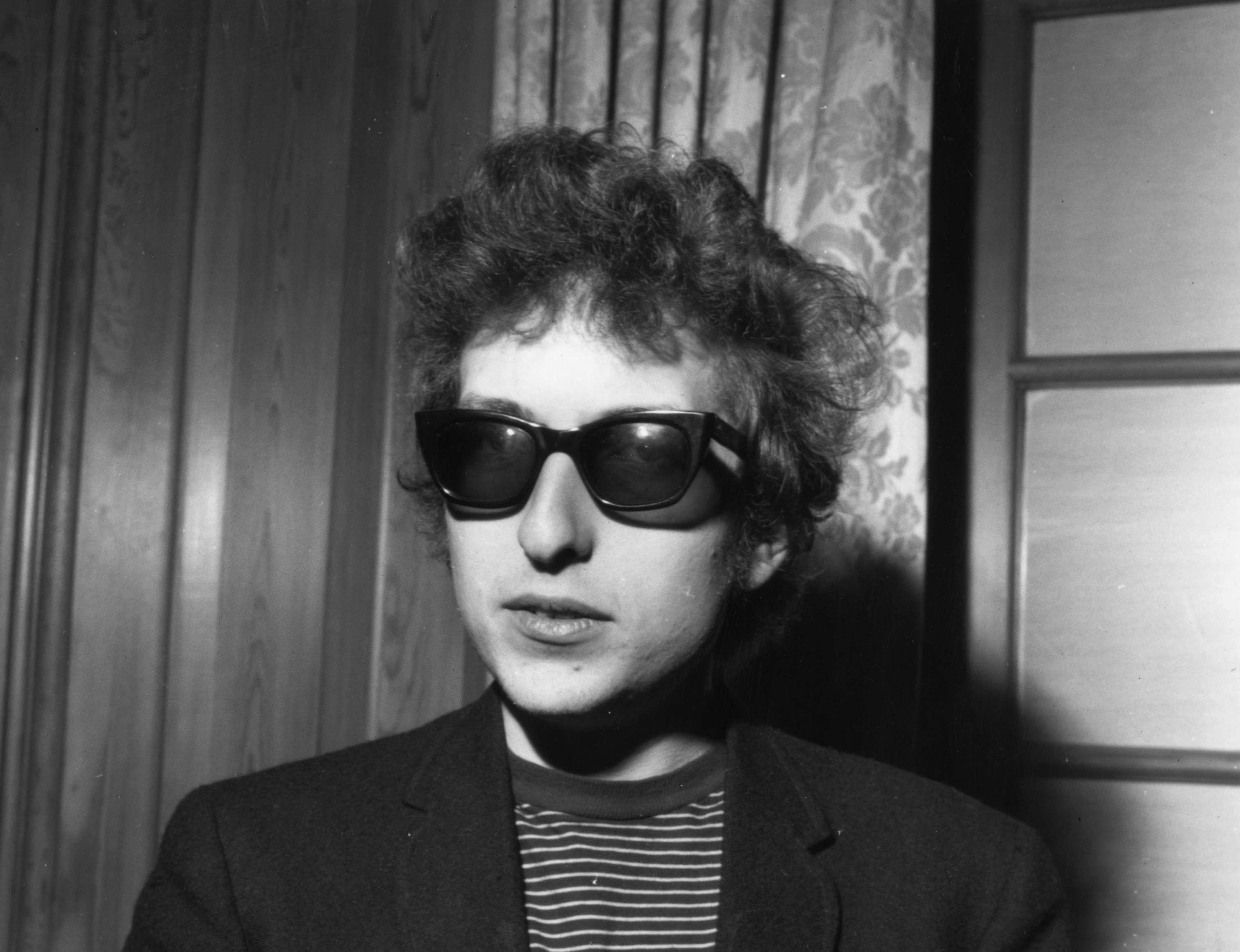 A black and white picture of Bob Dylan wearing sunglasses in front of a window.