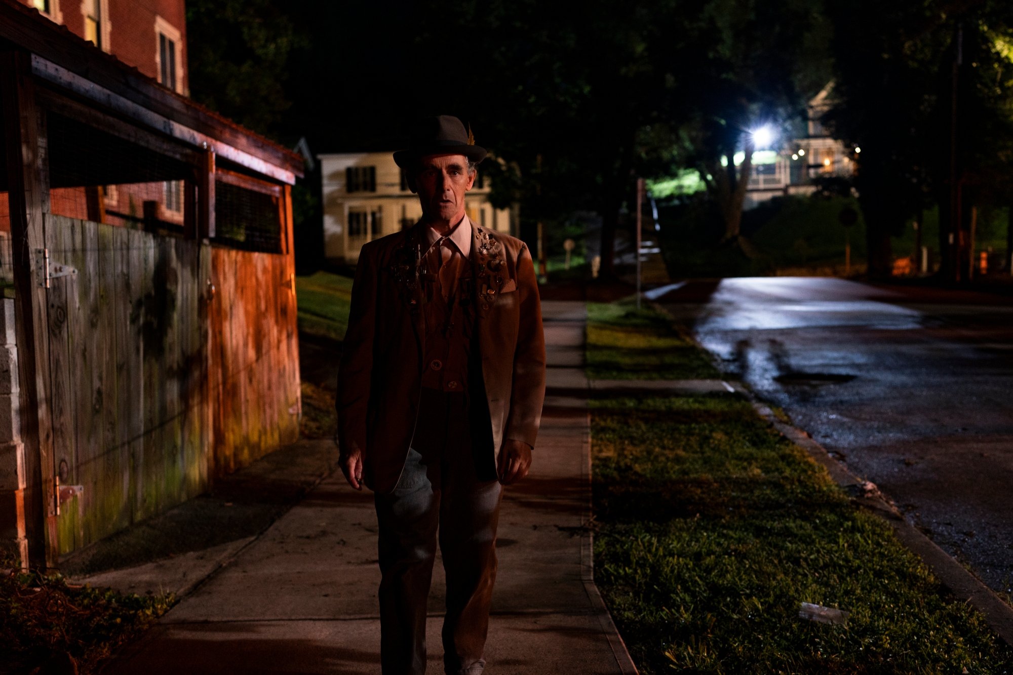 'Bones and All' Mark Rylance as Sully looking surprised standing on a dark street sidewalk. He's wearing a brown jacket, a collared shirt, and a hat.