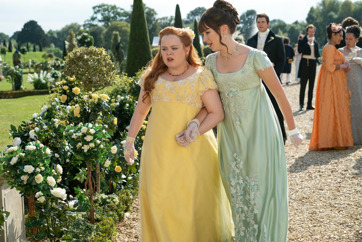 Nicola Coughlan as Penelope Featherington, and Claudia Jessie as Eloise Bridgerton chatting while walking in the gardensIn Bridgerton
