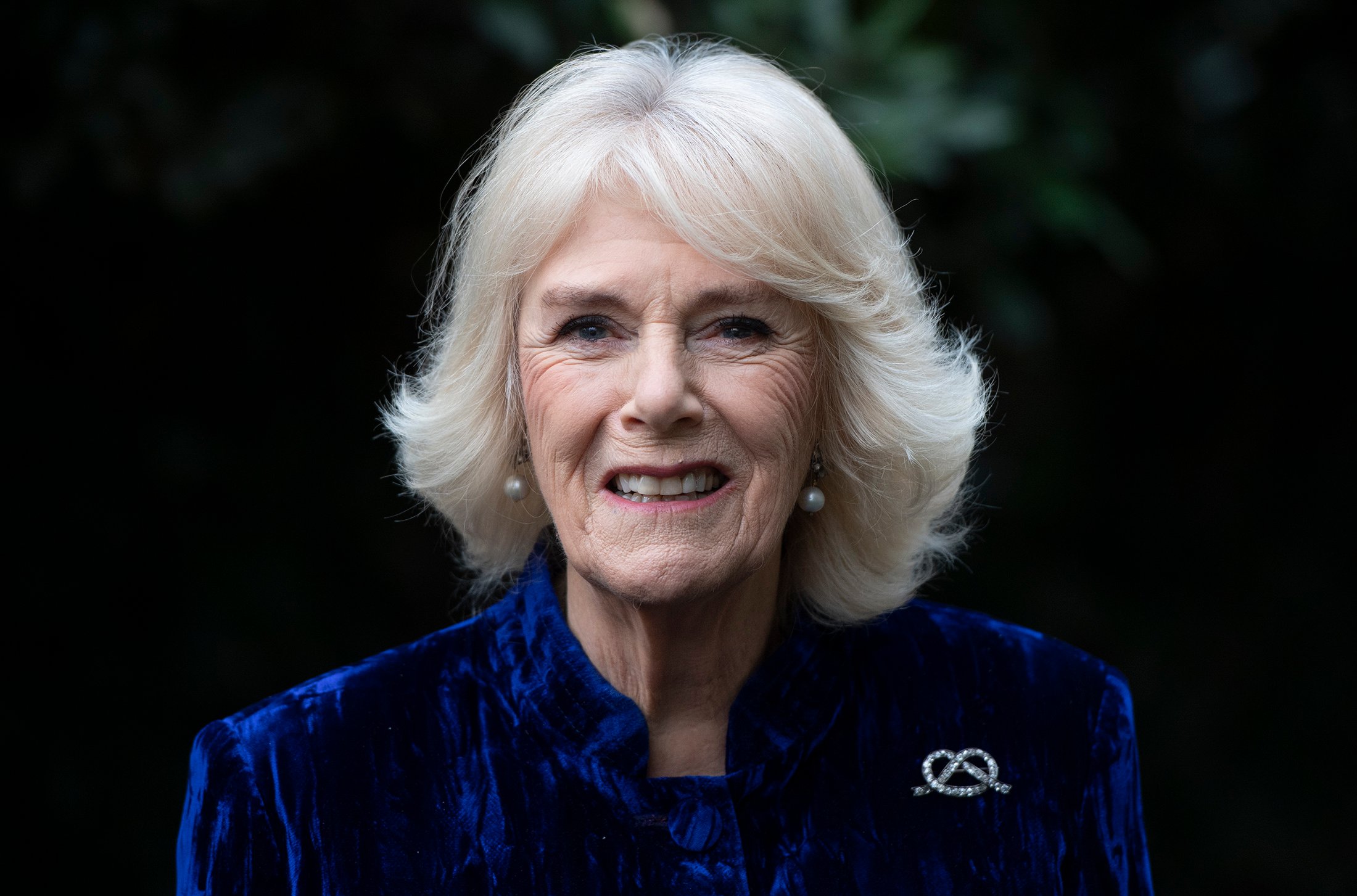 Camilla Parker Bowles, Queen Consort, smiles while wearing a royal blue velvet outfit.