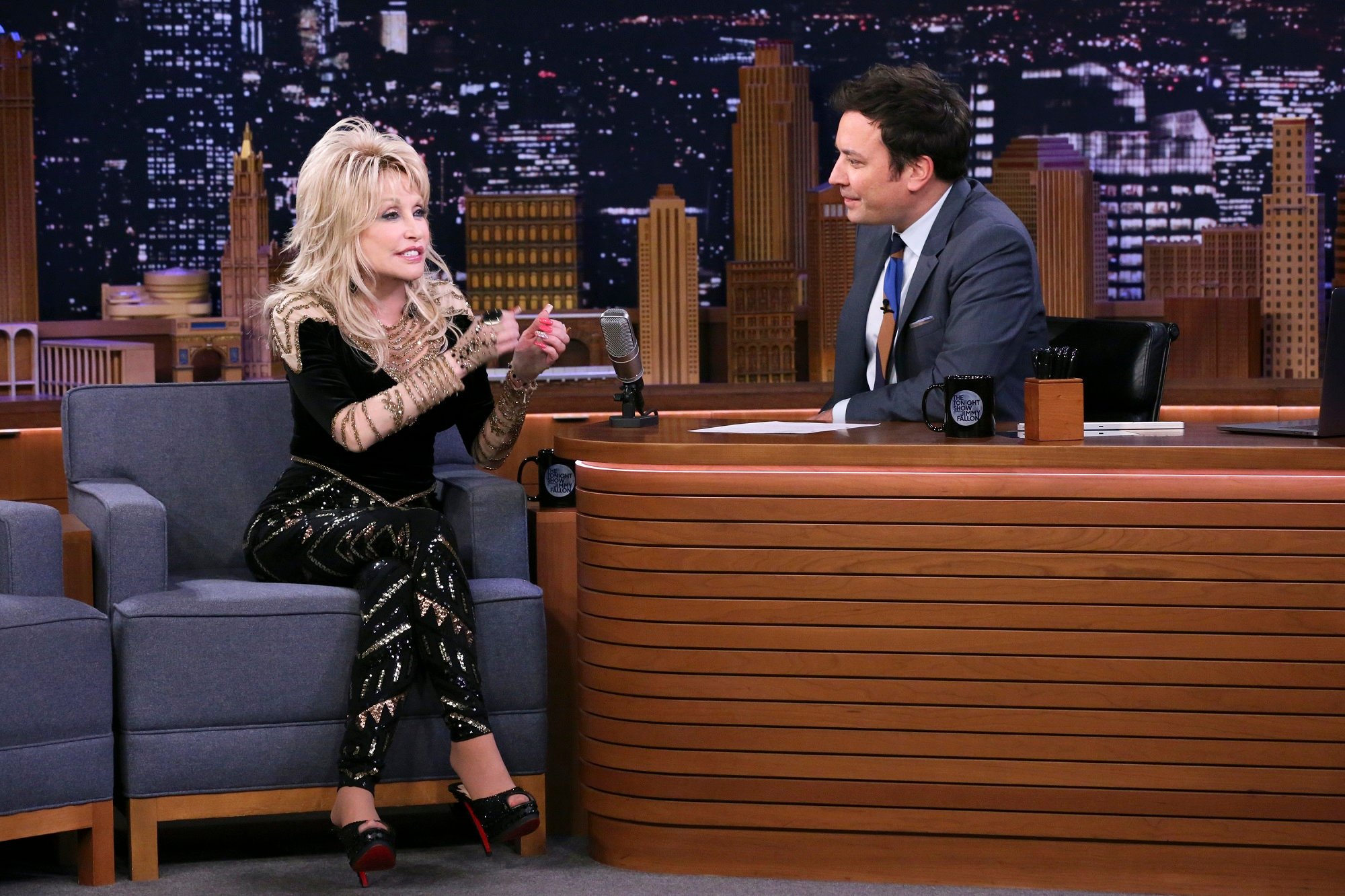 Dolly Parton sits on a gray chair and talks to Jimmy Fallon who is sitting behind a desk