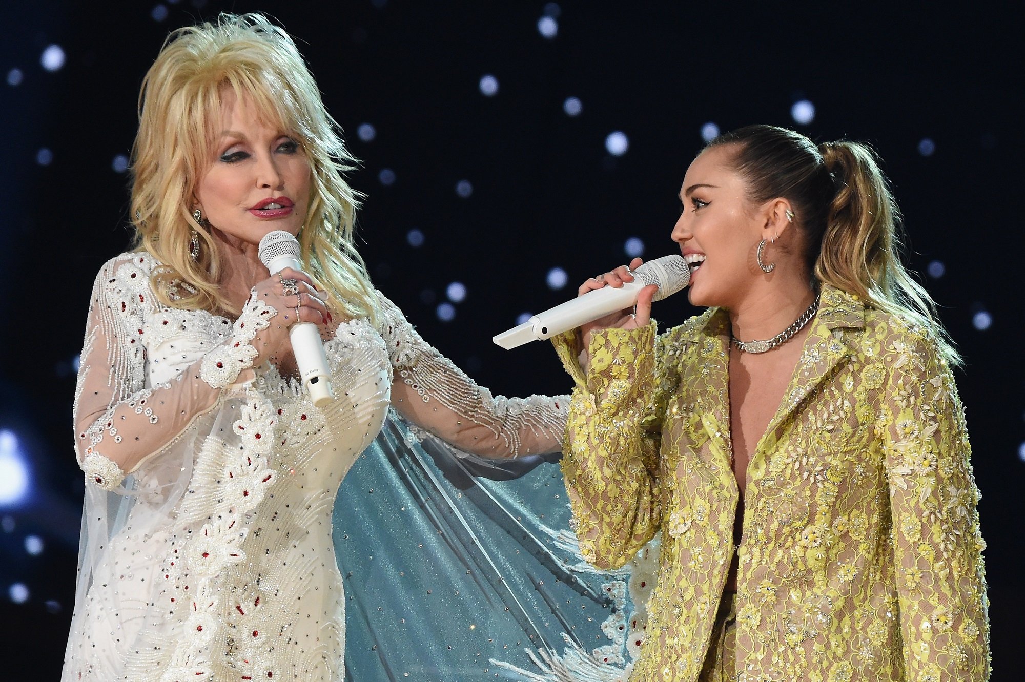 Dolly Parton and Miley Cyrus sing into white microphones side-by-side