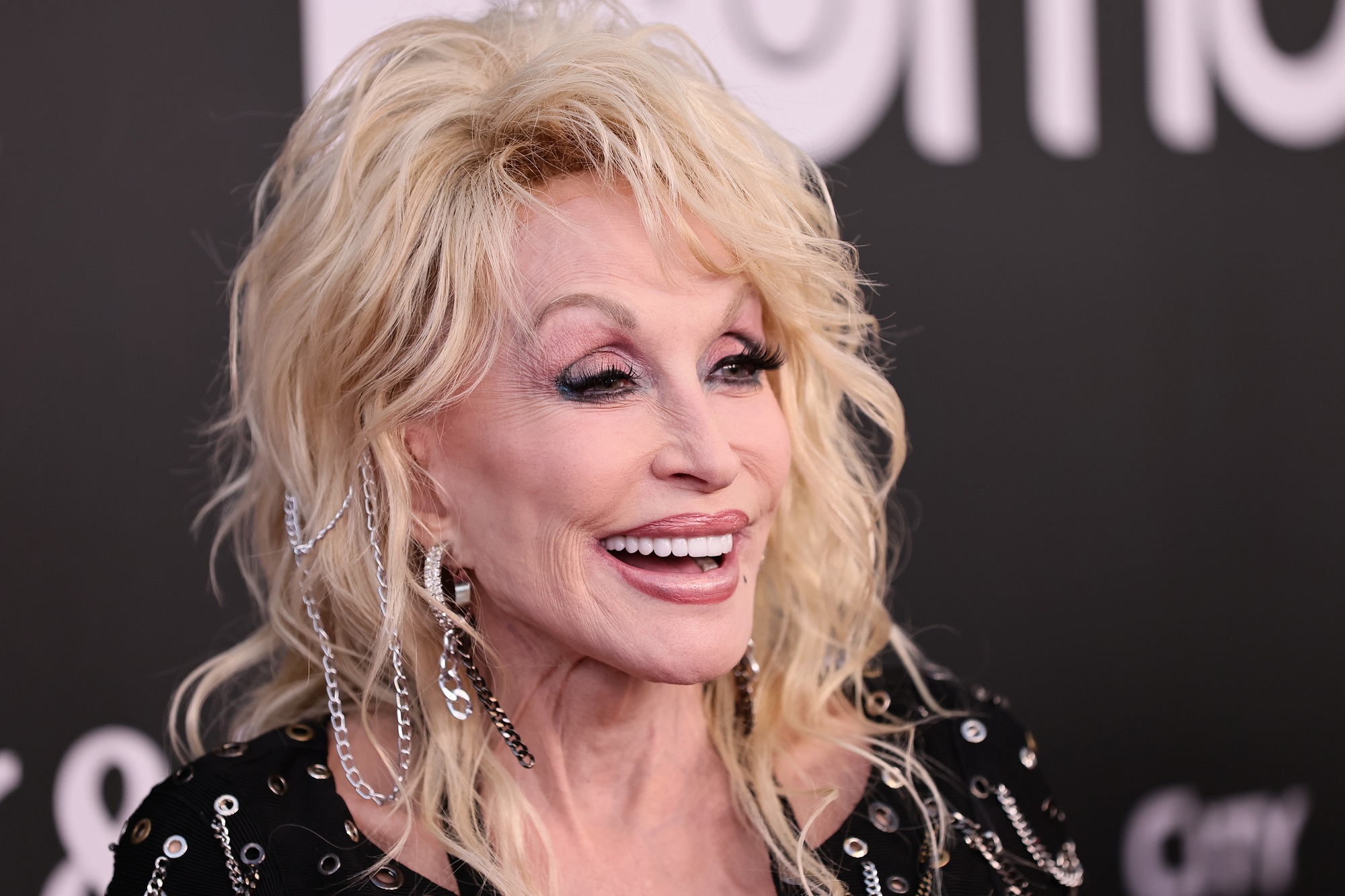 Dolly Parton smiles in front of a black backdrop with white writing
