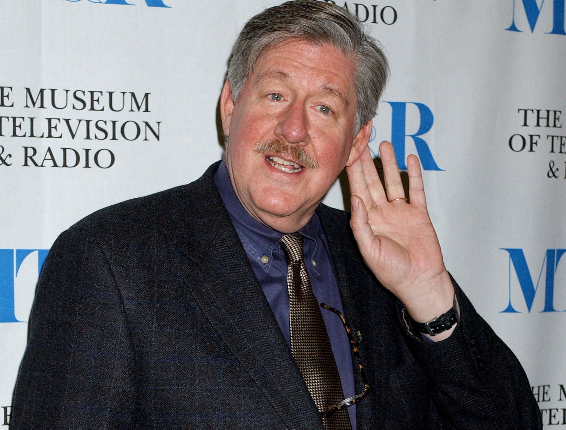 'Gilmore Girls' star Edward Herrmann, who criticized the real town Stars Hollow is based on. He's wearing a blue shirt, black suit, and holding his hand to his ear.