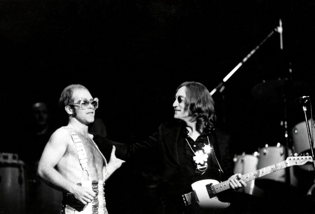 A black and white picture of Elton John and John Lennon onstage together. Lennon has a guitar.