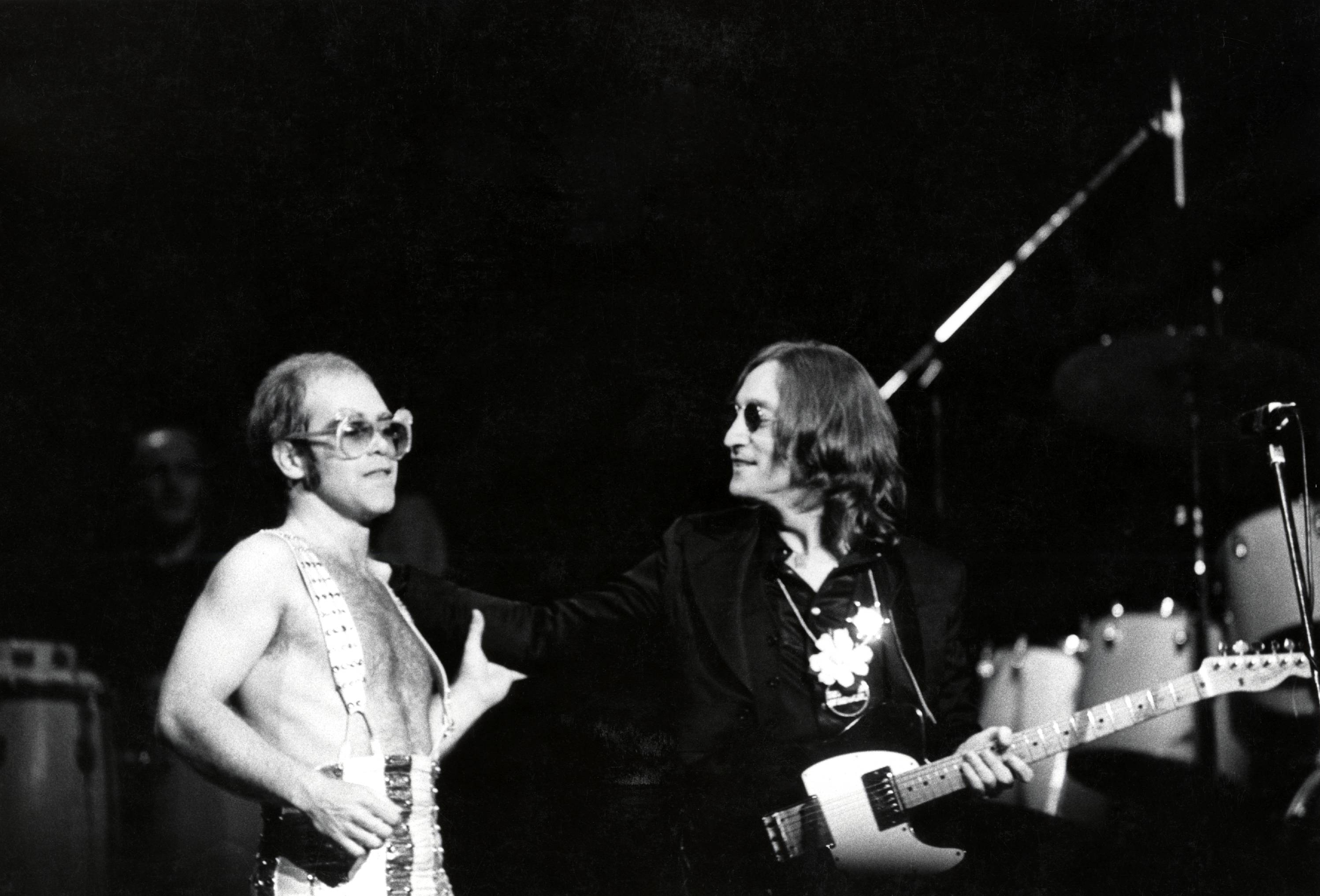 A black and white picture of Elton John and John Lennon on stage together. Lennon holds a guitar.