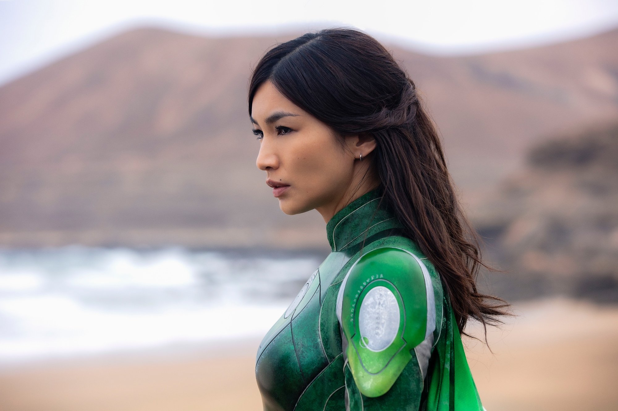 'Eternals' Gemma Chan as Sersi in MCU Phase 4 ranked list. She's wearing a green and silver suit, looking off into the distance on a beach with waves crashing in the background