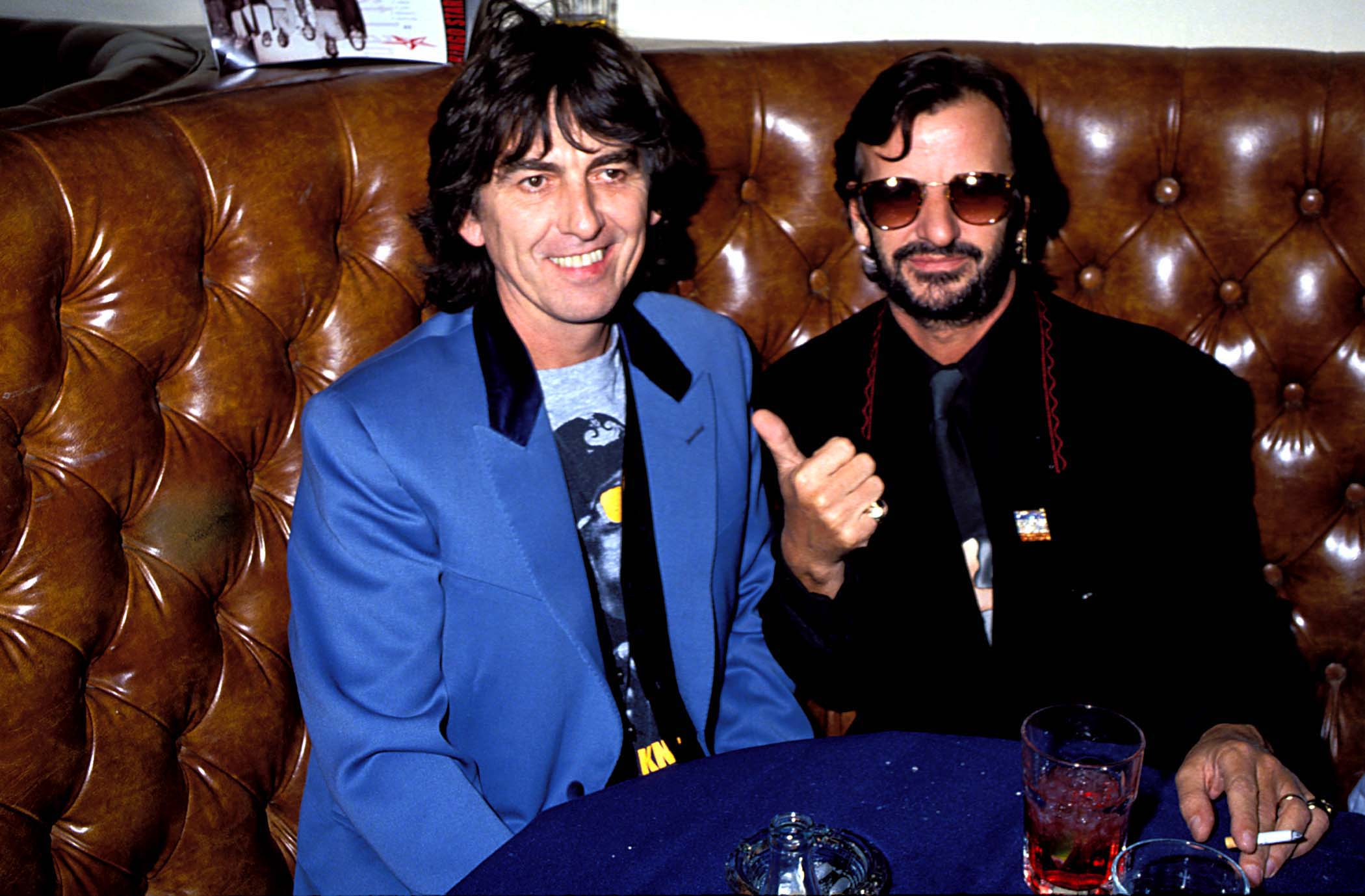 George Harrison and Ringo Starr sit at a table in a brown leather booth.