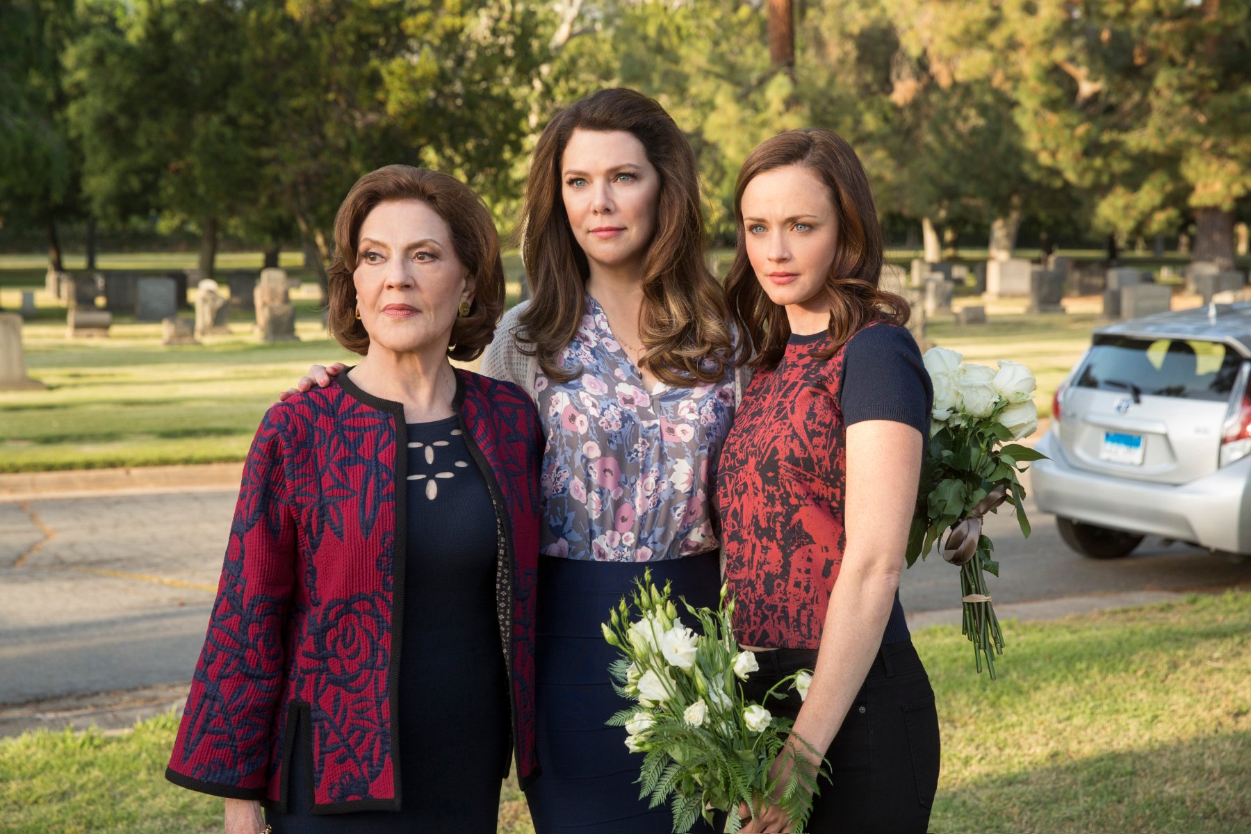 Kelly Bishop, Lauren Graham, and Alexis Bledel as Emily, Lorelai, and Rory in 'Gilmore Girls: A Year in the Life' for our article about lines from the original show. They're standing together outside, and Rory is holding white flowers.