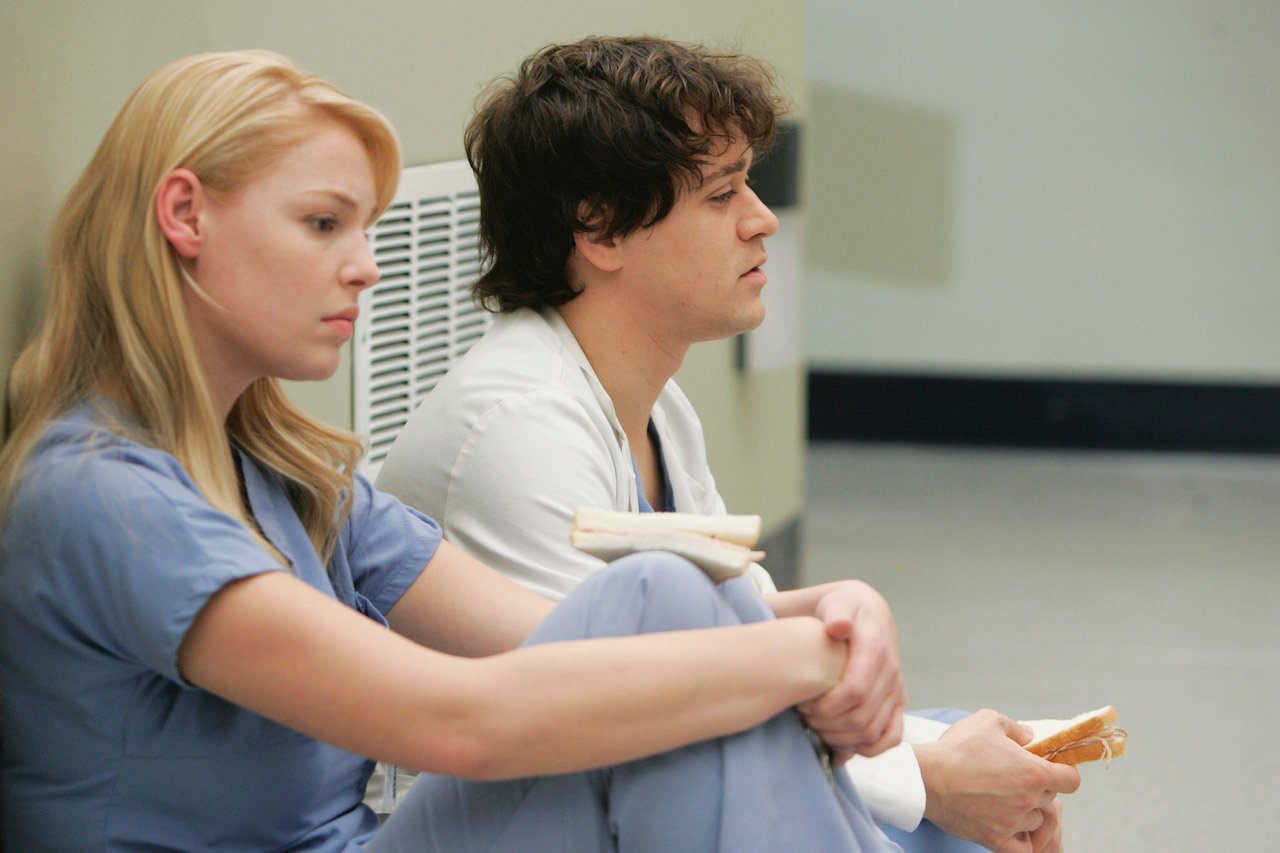 Katherine Heigl as Izzie Stevens and T.R. Knight as George O'Malley sit on the ground together in scrubs on 'Grey's Anatomy'.