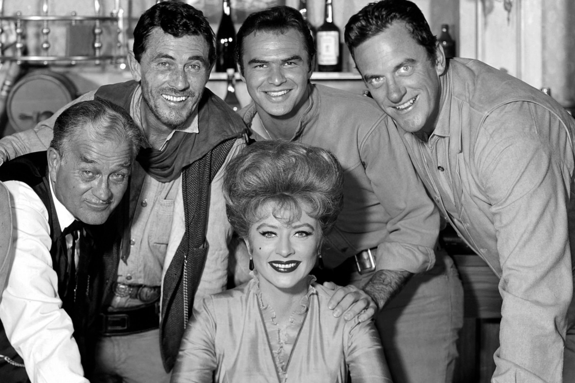 'Gunsmoke' Amanda Blake as Kitty Russell, Milburn Stone as Doc Adams, Ken Curtis as Festus Haggen, Burt Reynolds as Quint Asper, and James Arness as Matt Dillon in a black-and-white photograph. Blake is sitting down and her co-stars are all standing behind her smiling.