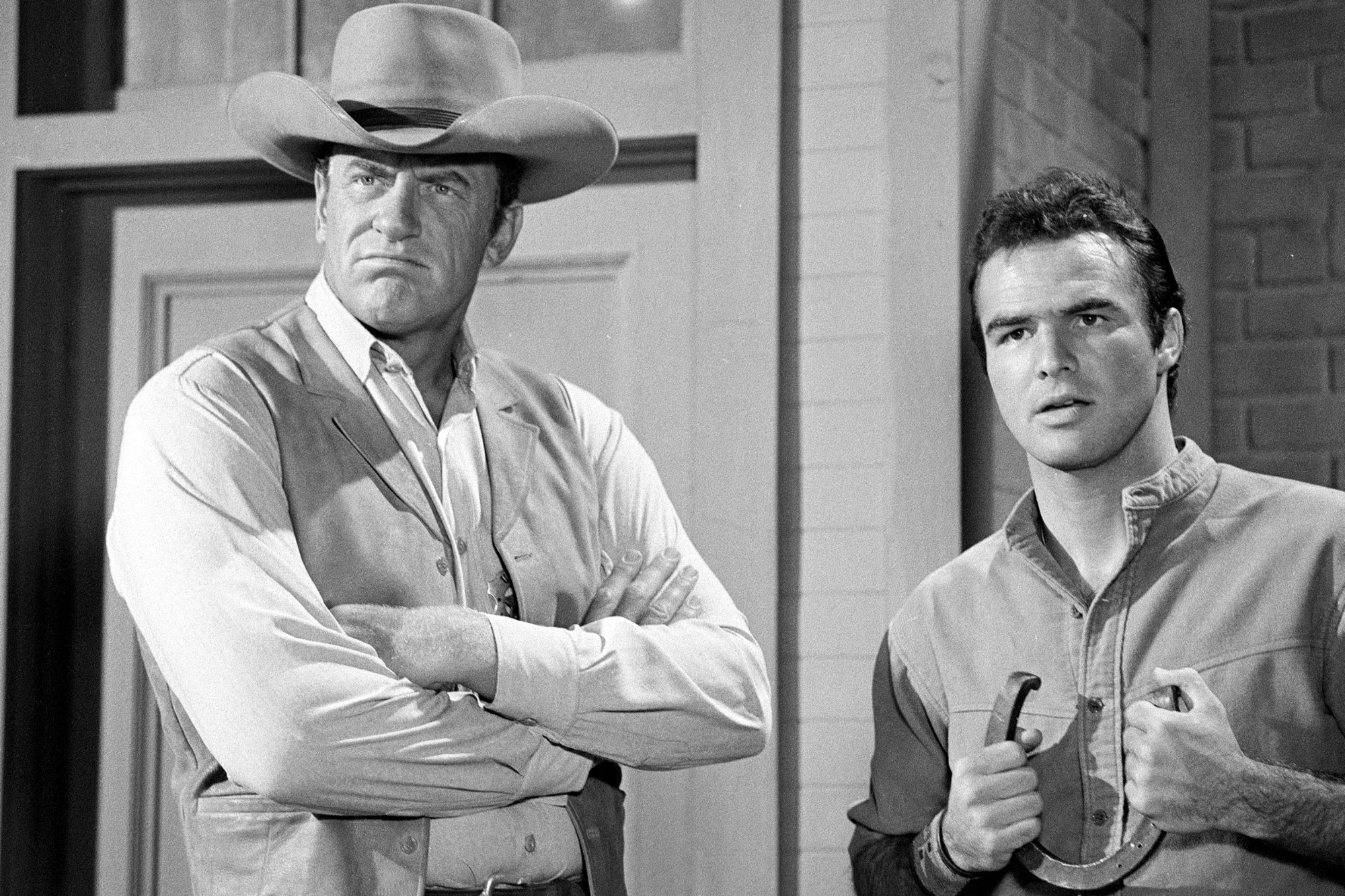 'Gunsmoke' James Arness as Matt Dillon and Burt Reynolds as Quint Asper. Arness has his arms crossed with a frown on his face. Reynolds is holding a horseshoe with his mouth slightly open.
