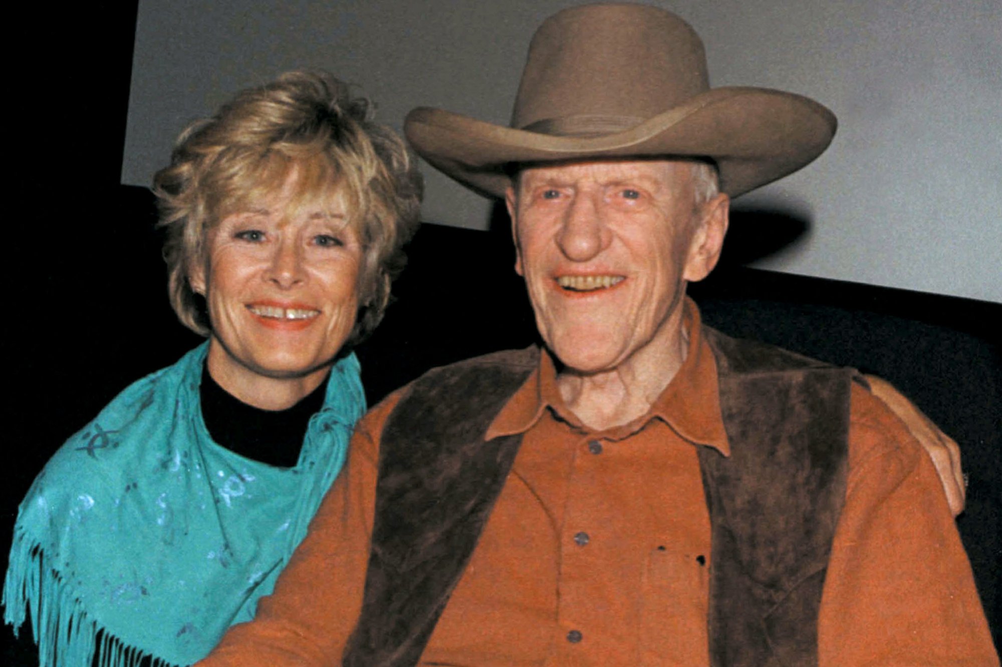 'Gunsmoke' actor James Arness and his wife, Janet, sitting next to each other. Arness is wearing a Western hat, vest, and collared shirt. Janet is wearing a blue scarf.