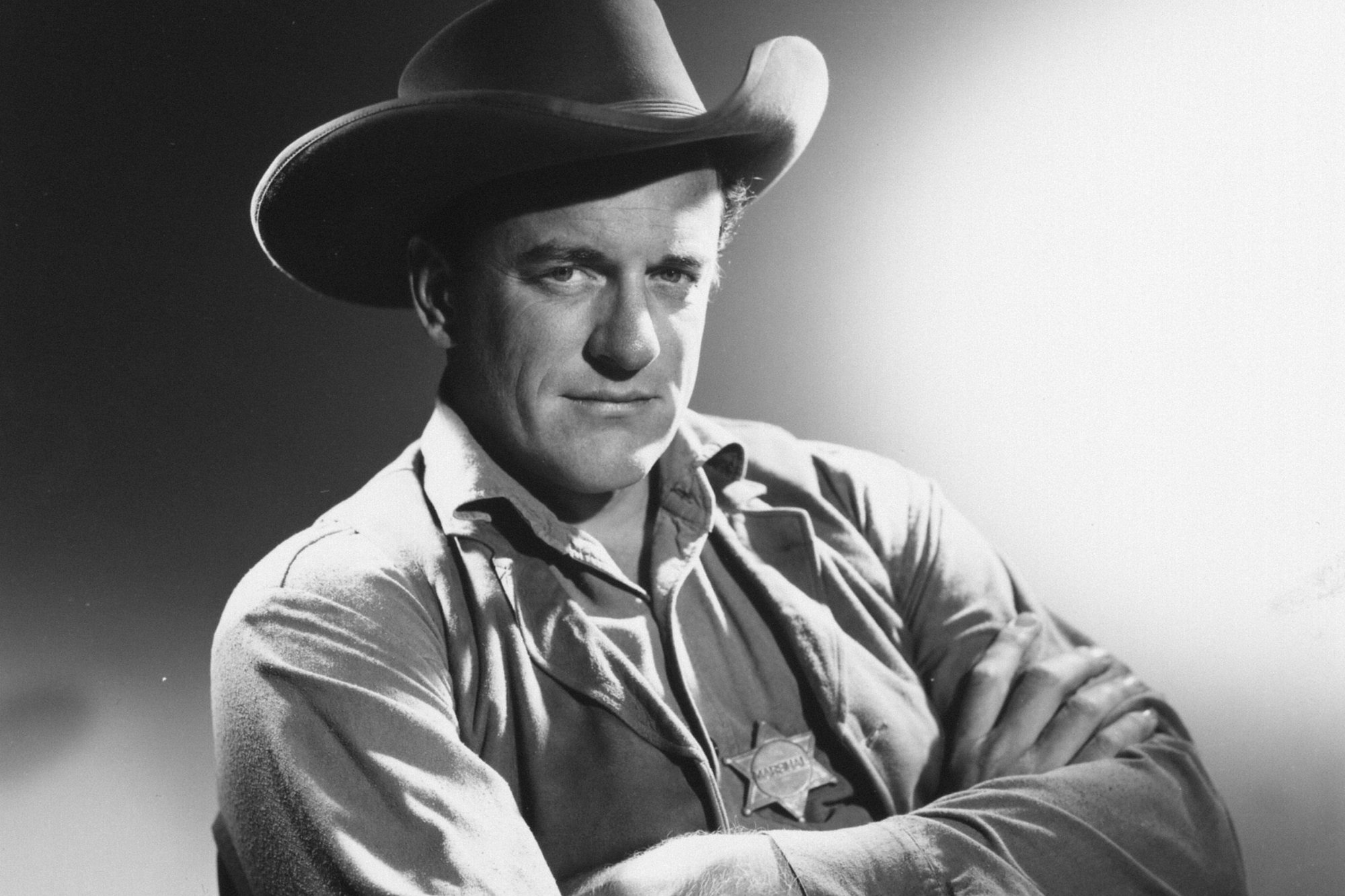 'Gunsmoke' actor James Arness as U.S. Marshal Matt Dillon with his arms crossed in a black-and-white picture. He has his marshal badge on his vest and is wearing a Western hat.