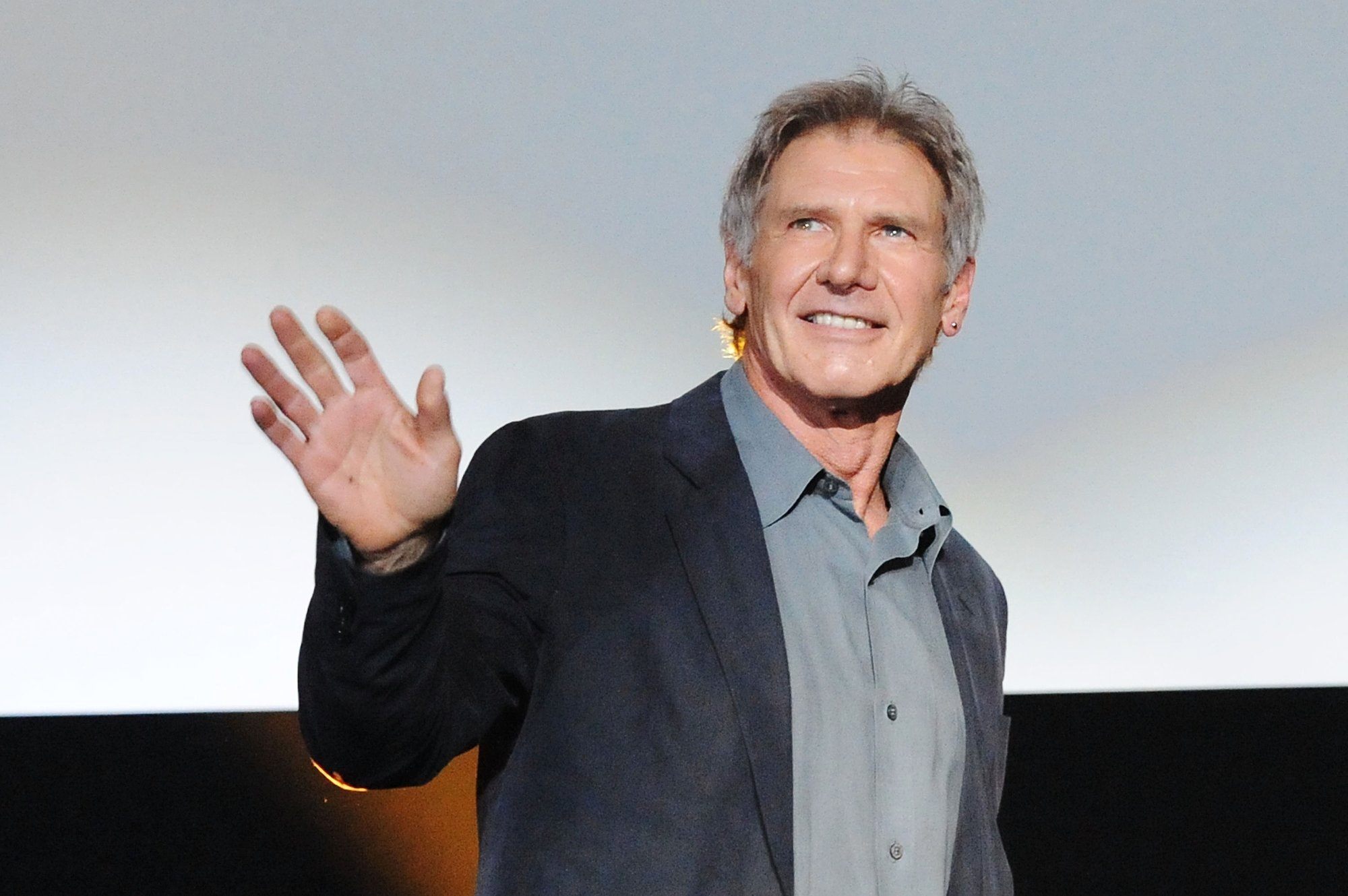 'Gunsmoke' guest star Harrison Ford. He's holding up his hand waving in front of a silver screen, wearing a collared shirt and suit jacket
