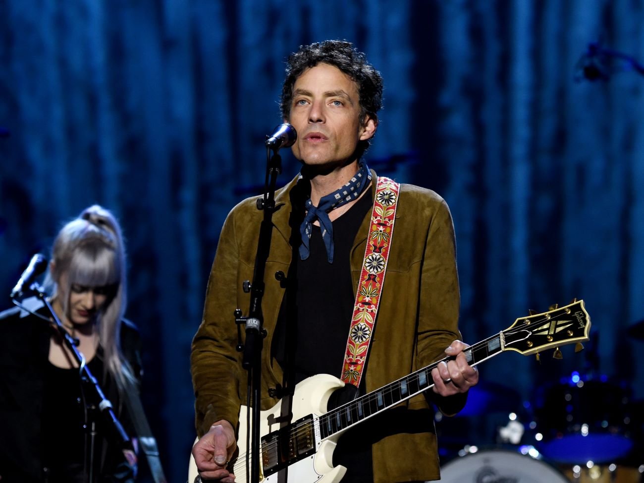 Jakob Dylan plays guitar and stands in front of a microphone. 