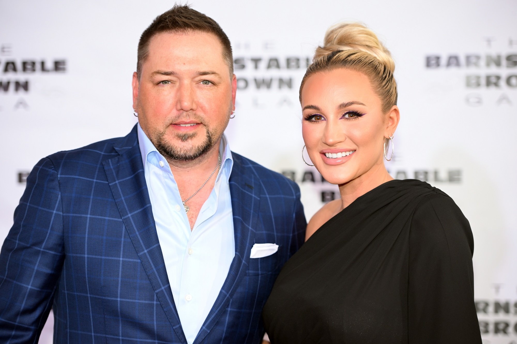 Jason Aldean and his wife Brittany Aldean smile at the camera in front of a white backdrop