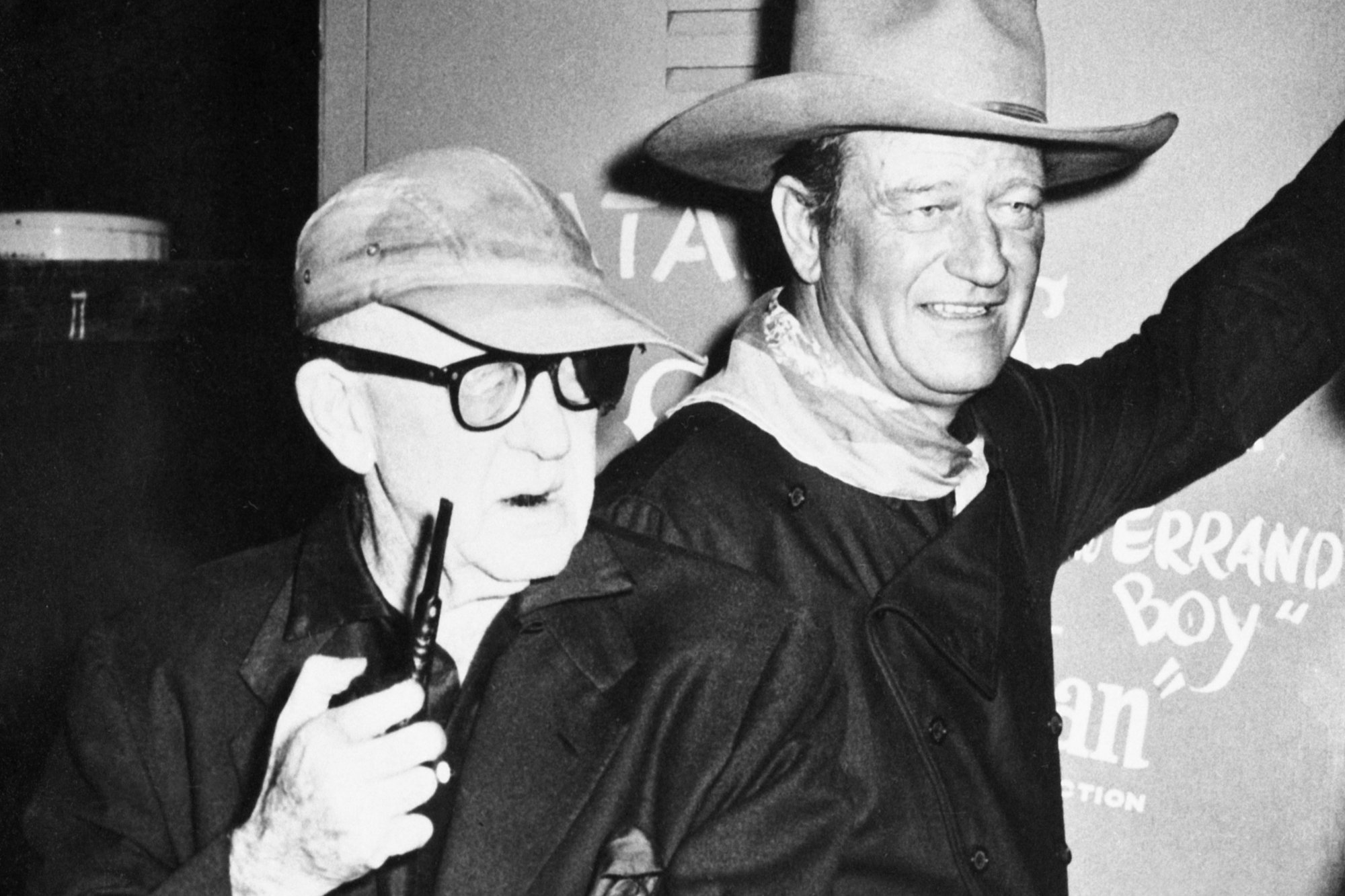John Ford and John Wayne standing next to each other in a black-and-white photograph. Wayne is in-costume to start filming 'The Man Who Shot Liberty Valance'