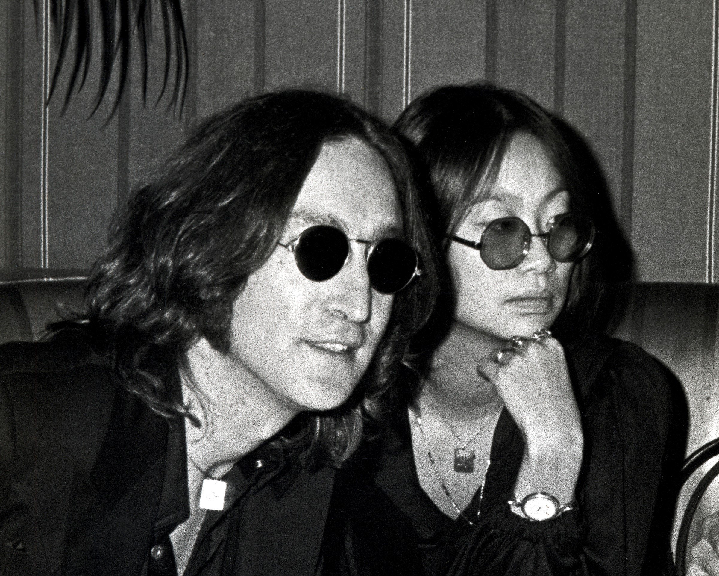A black and white picture of John Lennon and May Pang sitting together and wearing sunglasses.