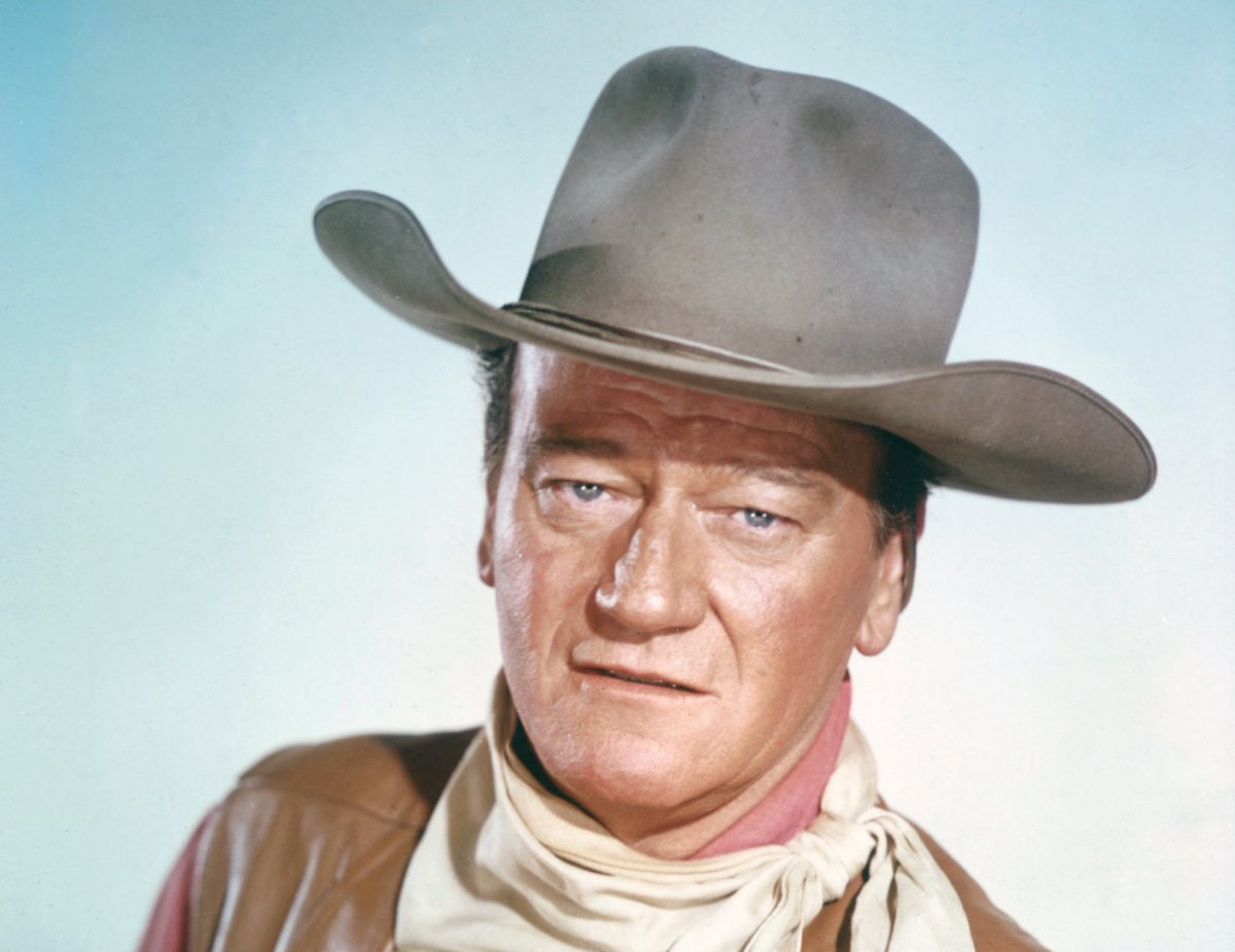 John Wayne (1907 - 1979), US actor wearing a tan leather waistcoat, a pink shirt and a white neckerchief, in a studio portrait, against a light blue background, circa 1970