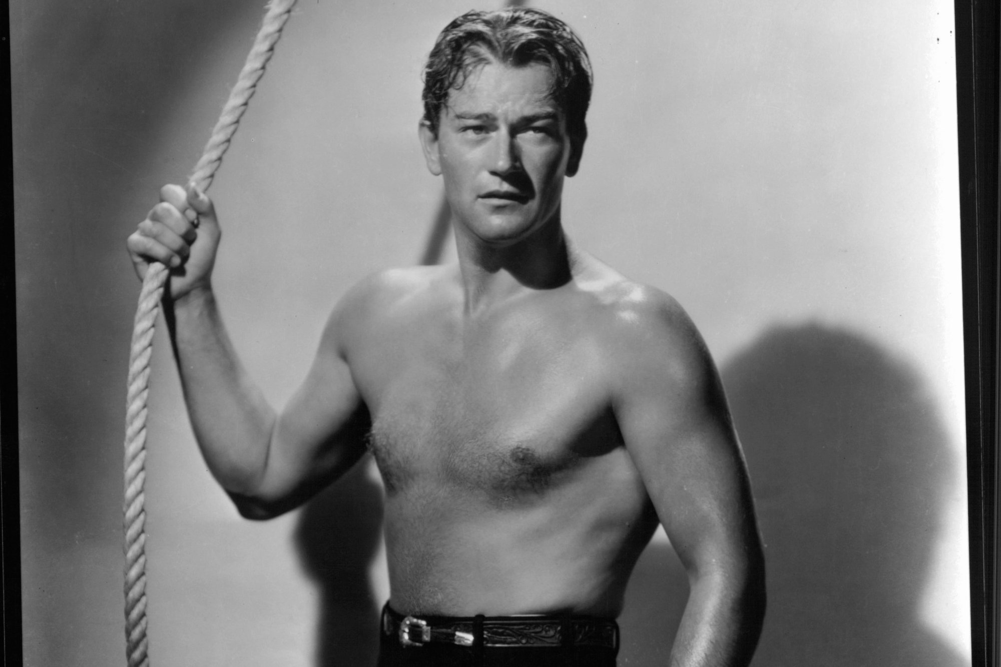 John Wayne, whose works have been called queer 'erotic spectacle.' He's shirtless in a black-and-white picture, holding onto a rope.