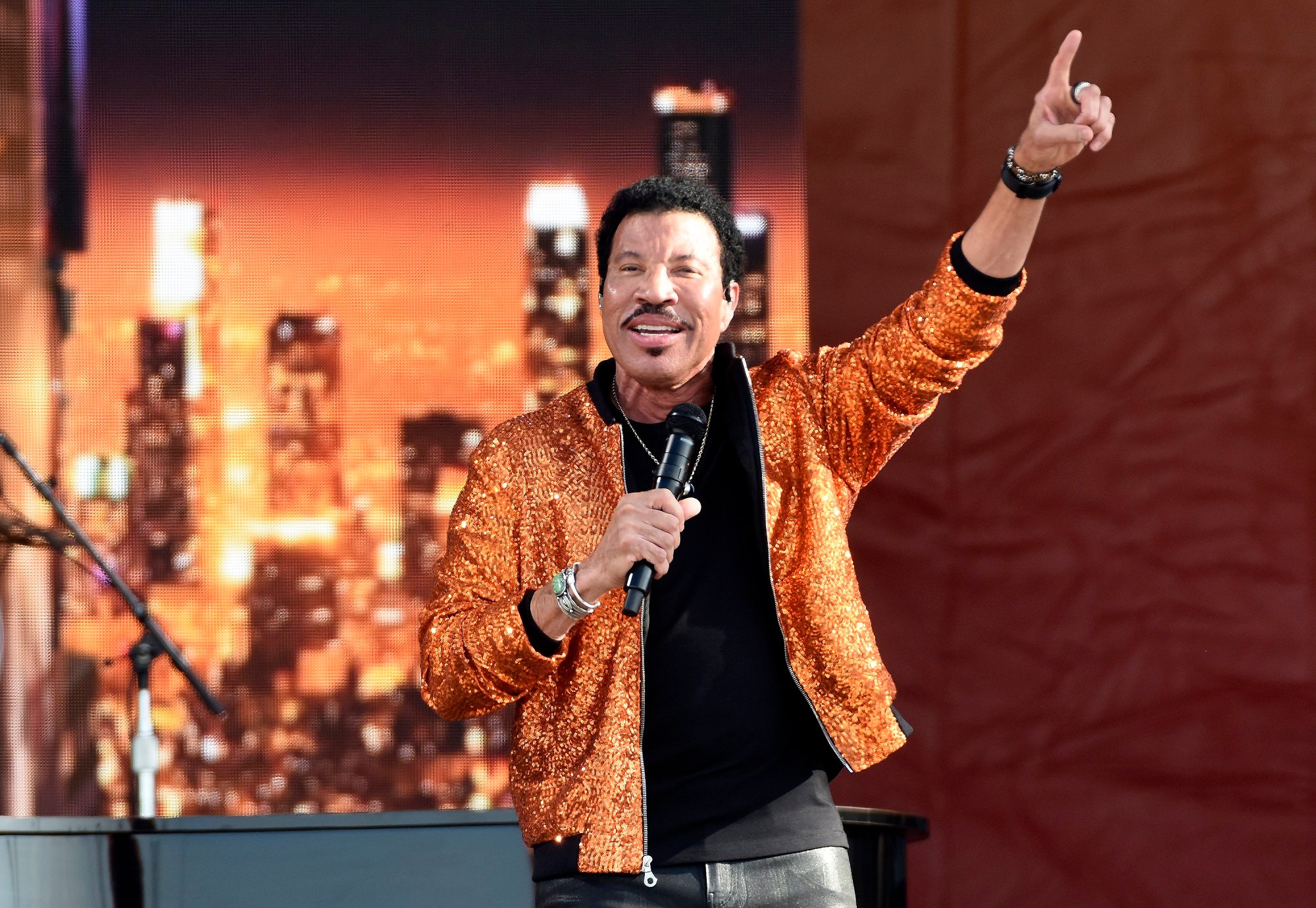 Lionel Richie stands on stage in an orange shimmery jacket
