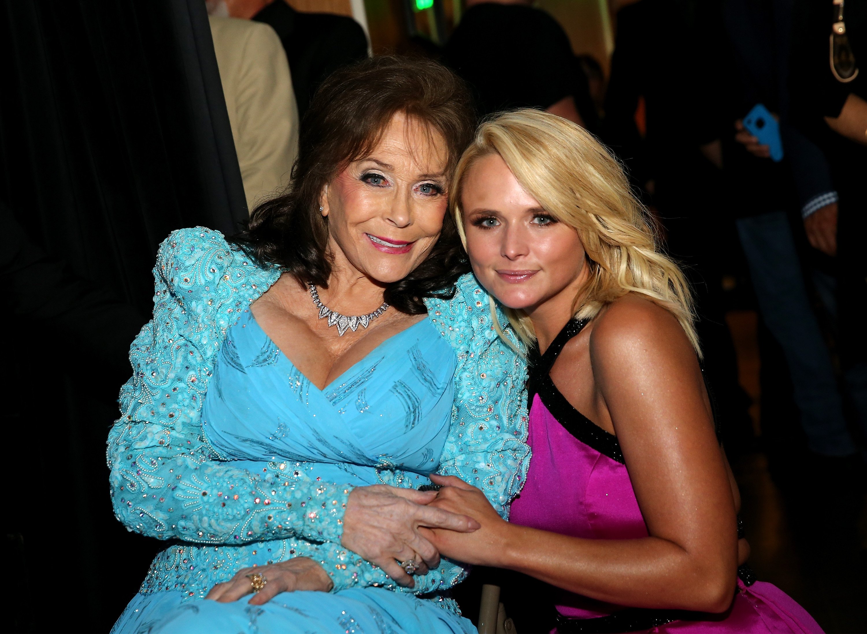 Loretta Lynn and Miranda Lambert sit beside each other holding hands while looking at the camera