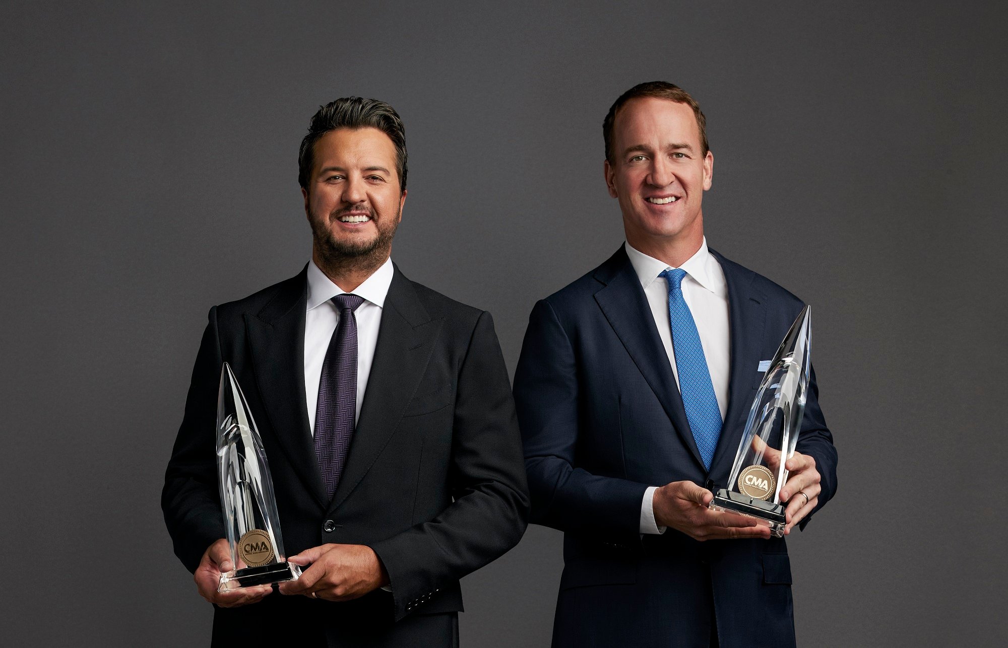 Luke Bryan and Peyton Manning pose with CMA trophies in front of a gray backdrop