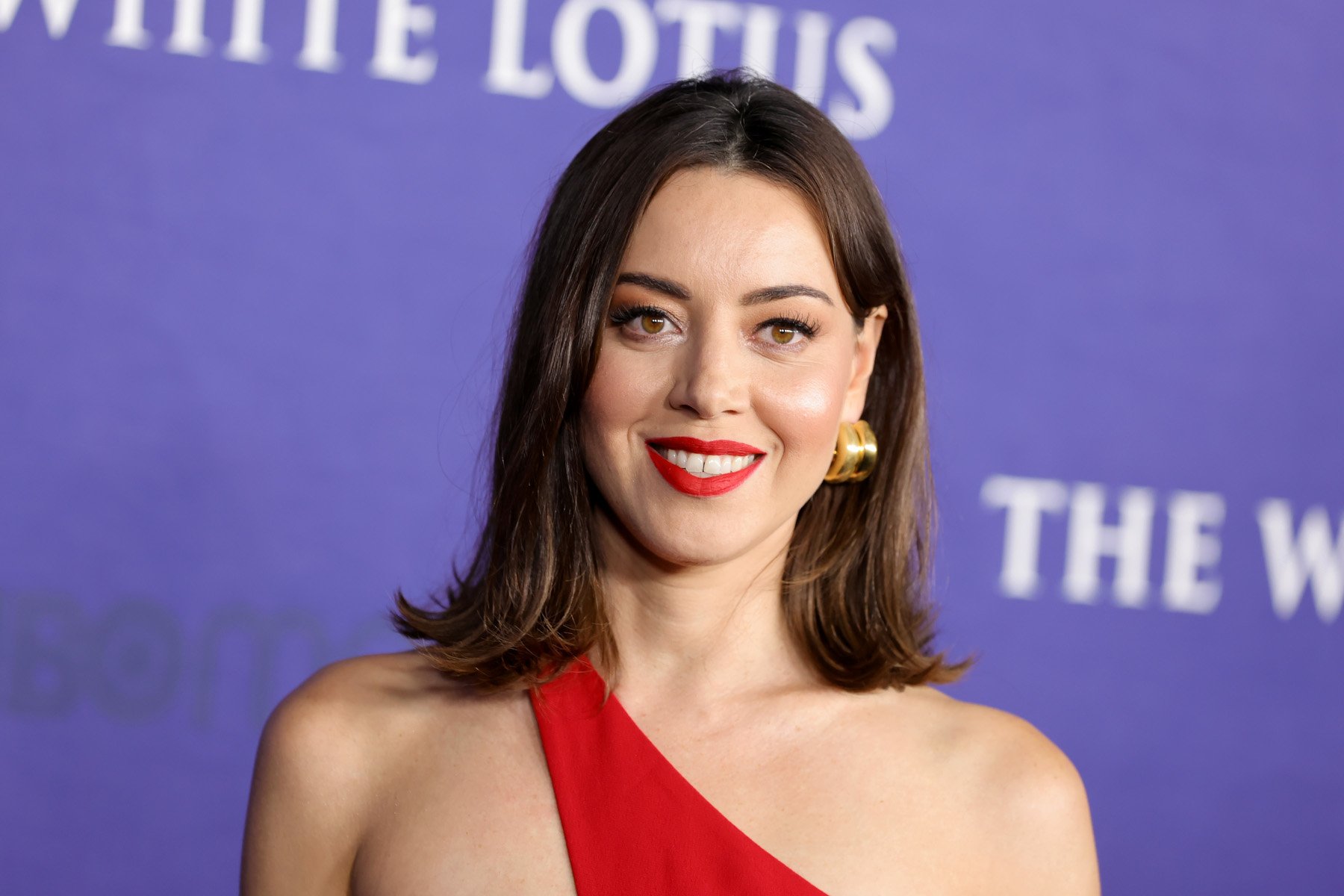 'Parks and Rec' cast member Aubrey Plaza. She's standing in front of a 'White Lotus' wall and has a red, one-strap dress on.