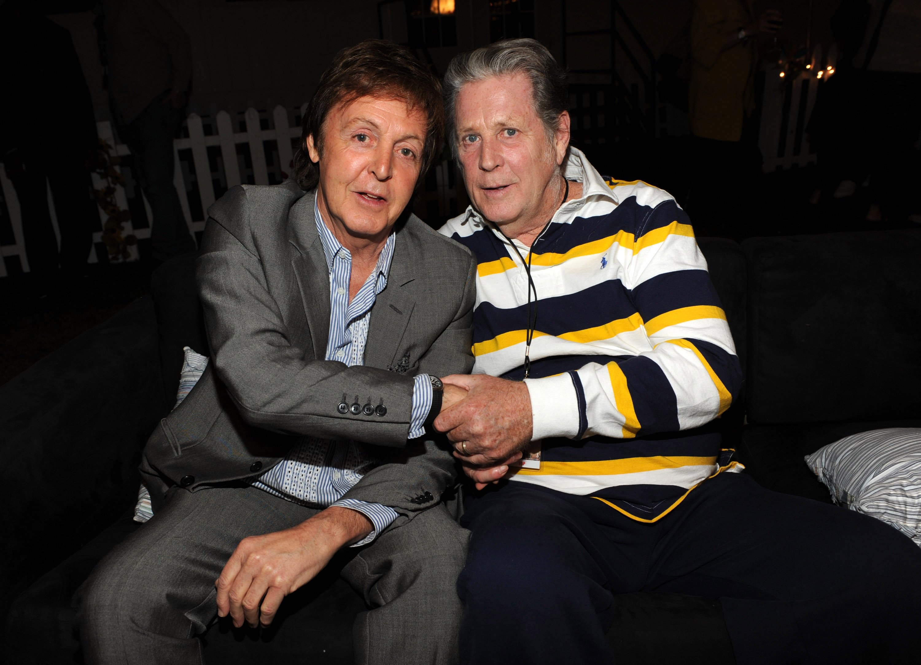 Paul McCartney and Brian Wilson sit next to each other and shake hands.