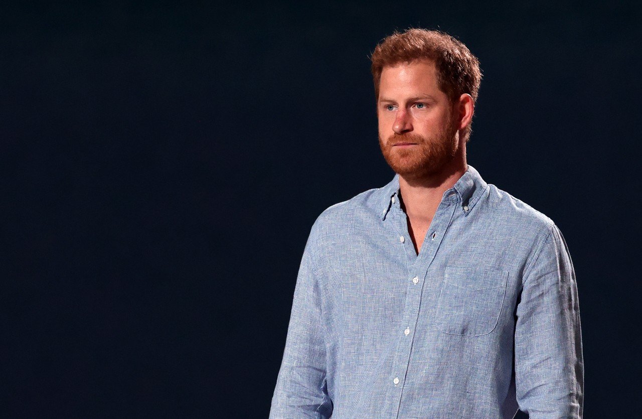 Prince Harry stands in front of a black background and wears a jean shirt.