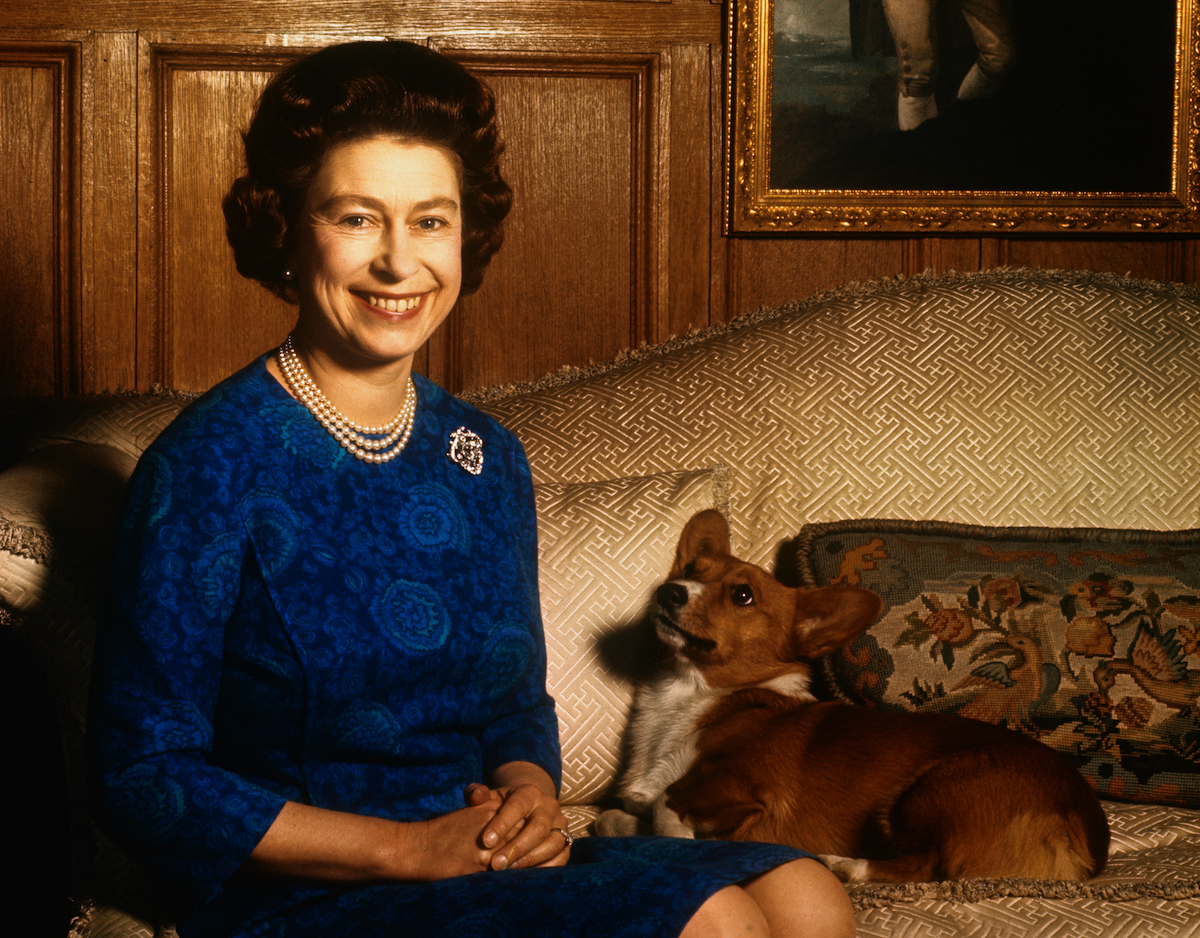 Queen Elizabeth, who, according to a dog trainer, said Prince Philip already told her she had too many dogs, wears a blue dress and smiles sitting with a corgi on a couch