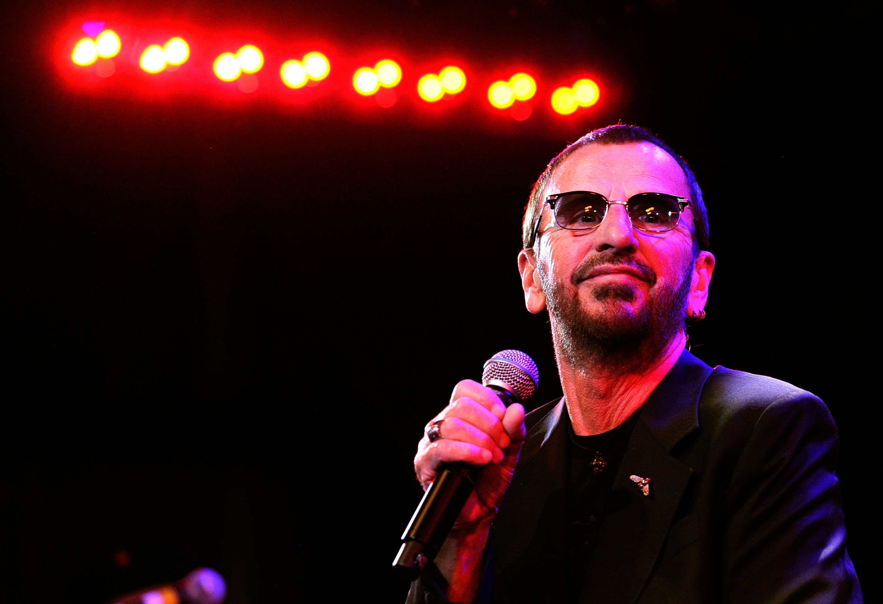 Ringo Starr wears sunglasses and holds a microphone.
