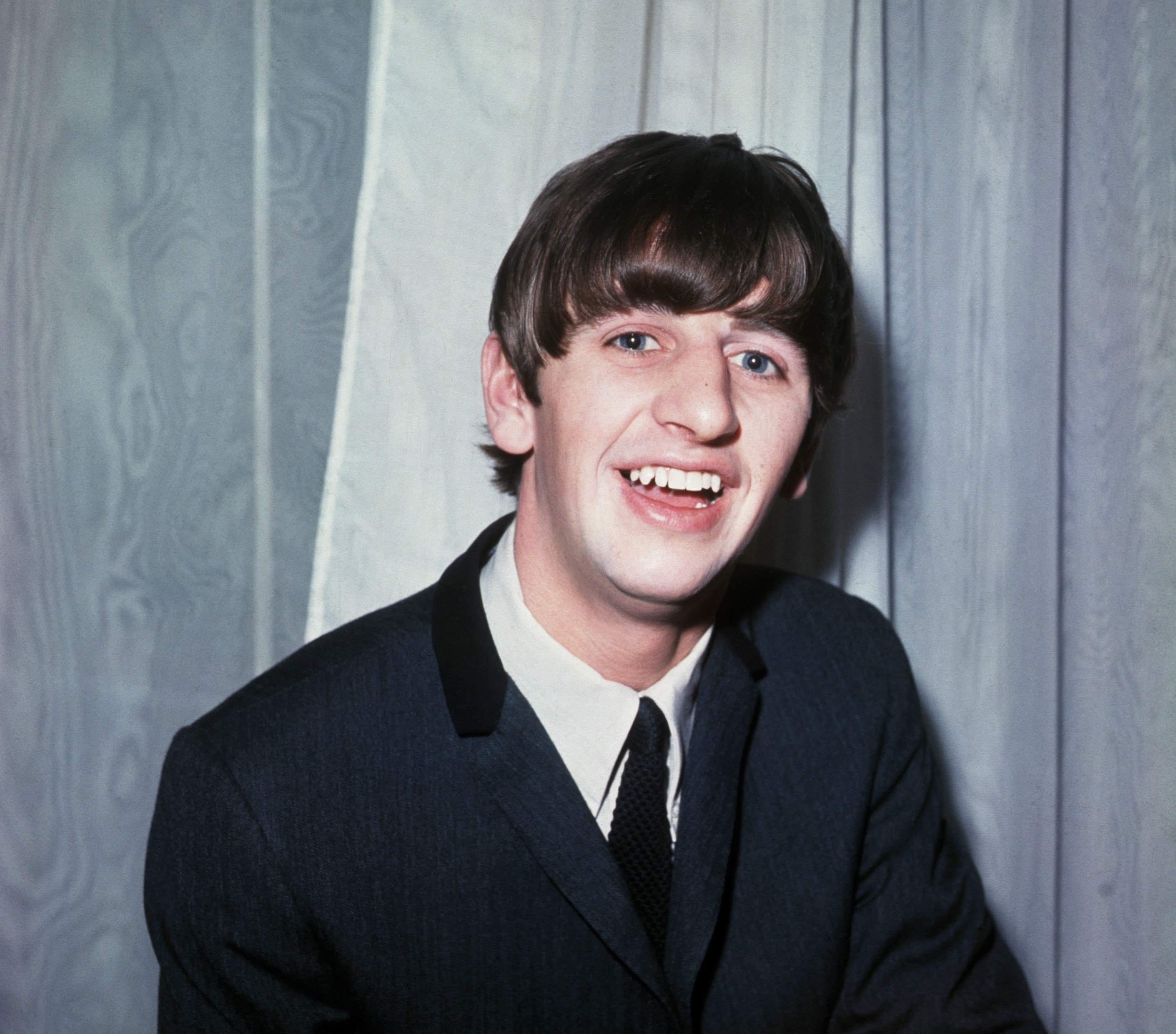 Ringo Starr wears a suit and sits in front of a gray background.