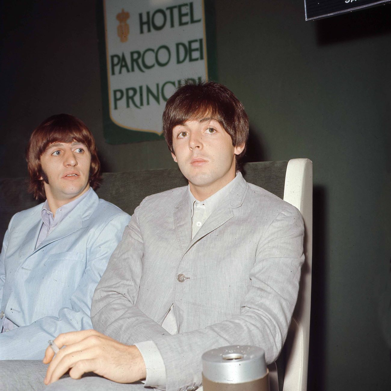 Ringo Starr wears a blue suit and sits next to Paul McCartney, who wears a gray suit. 
