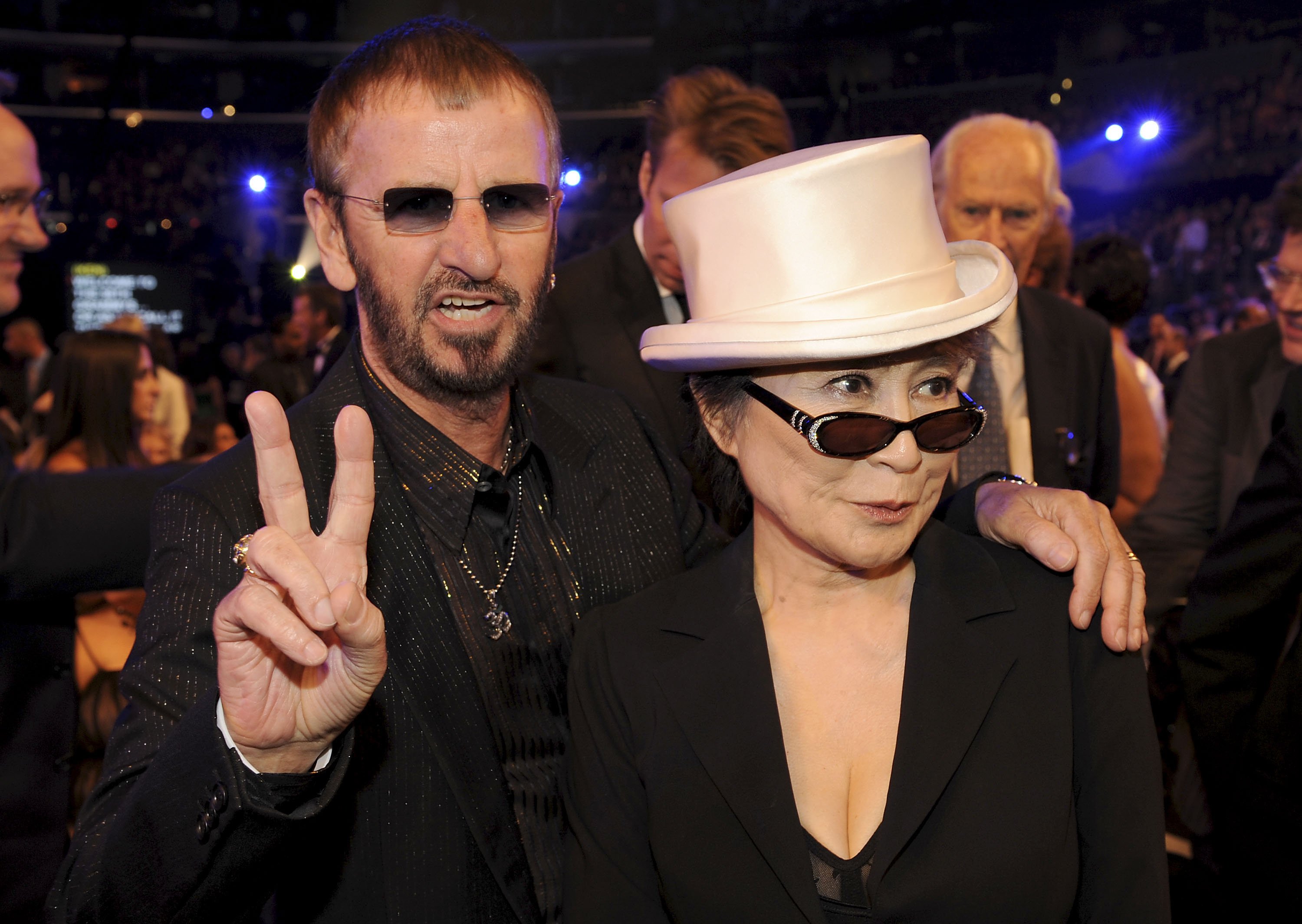 Ringo Starr and Yoko Ono wear black. Starr holds up a peace sign.
