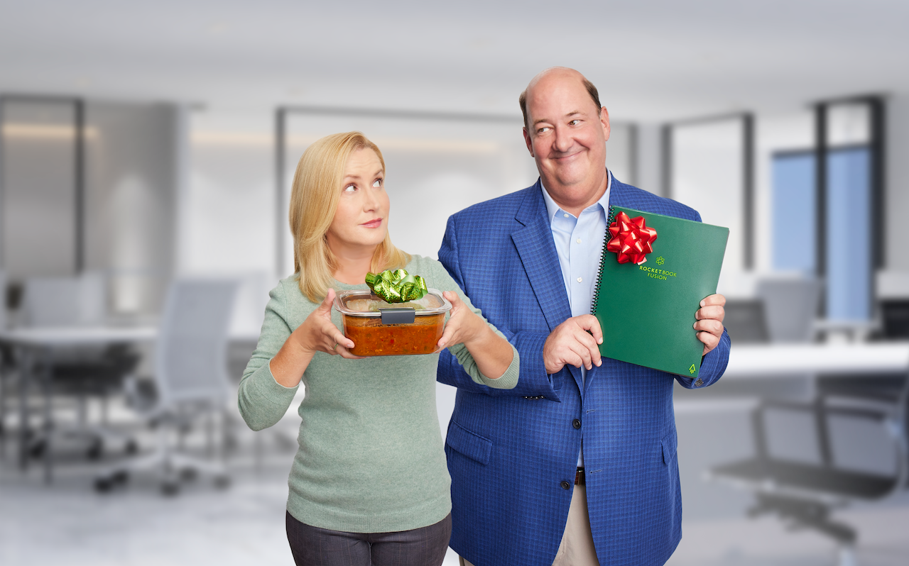 Angela Kinsey holds a bowl of chili standing next to Brian Baumgartner who holds up a Rocketbook.