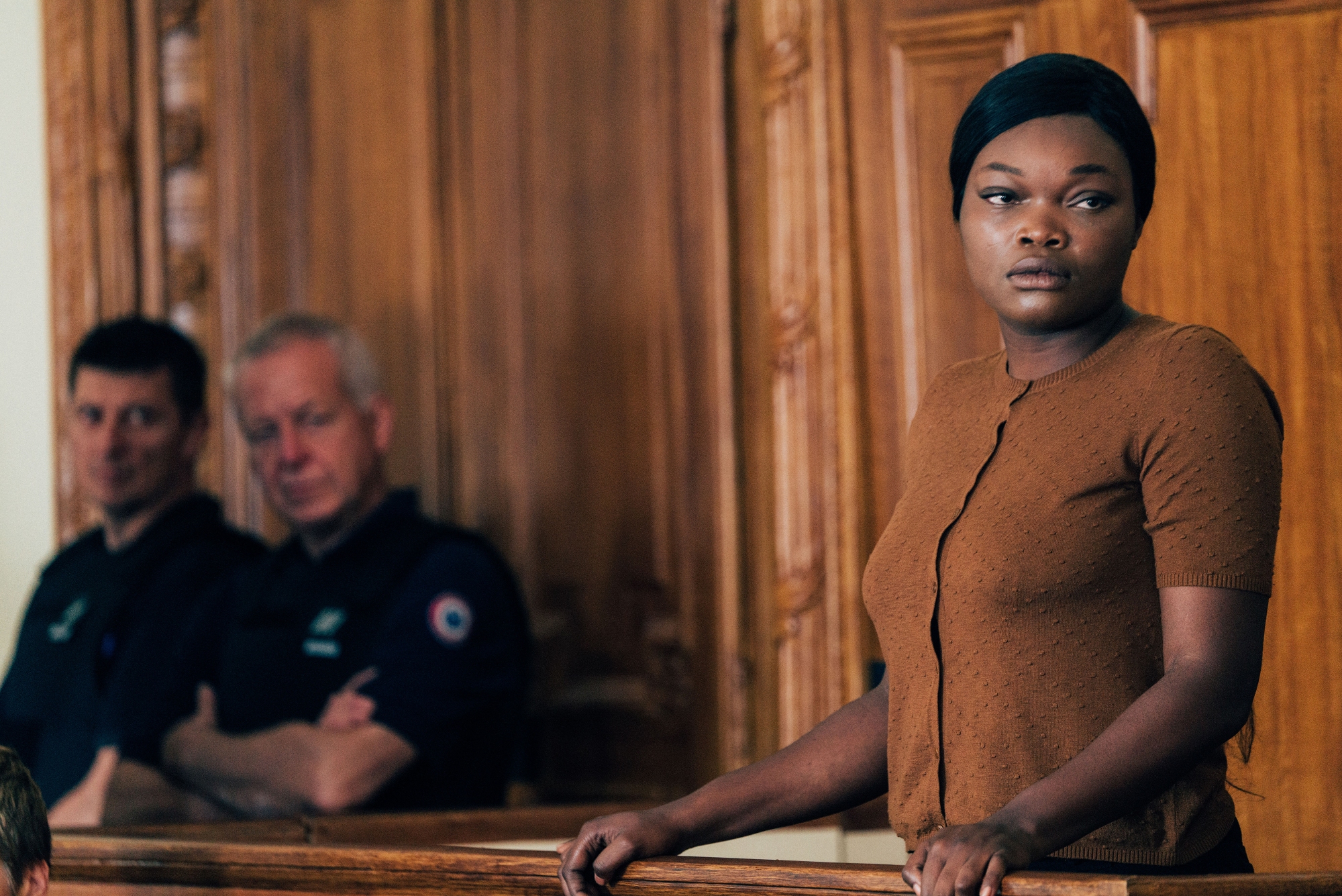 'Saint Omer' Guslagie Malanga as Laurence Coly standing at the court stand during a trial looking to the side. She's wearing a brown top. Two security guards look at her from the corner of the room.