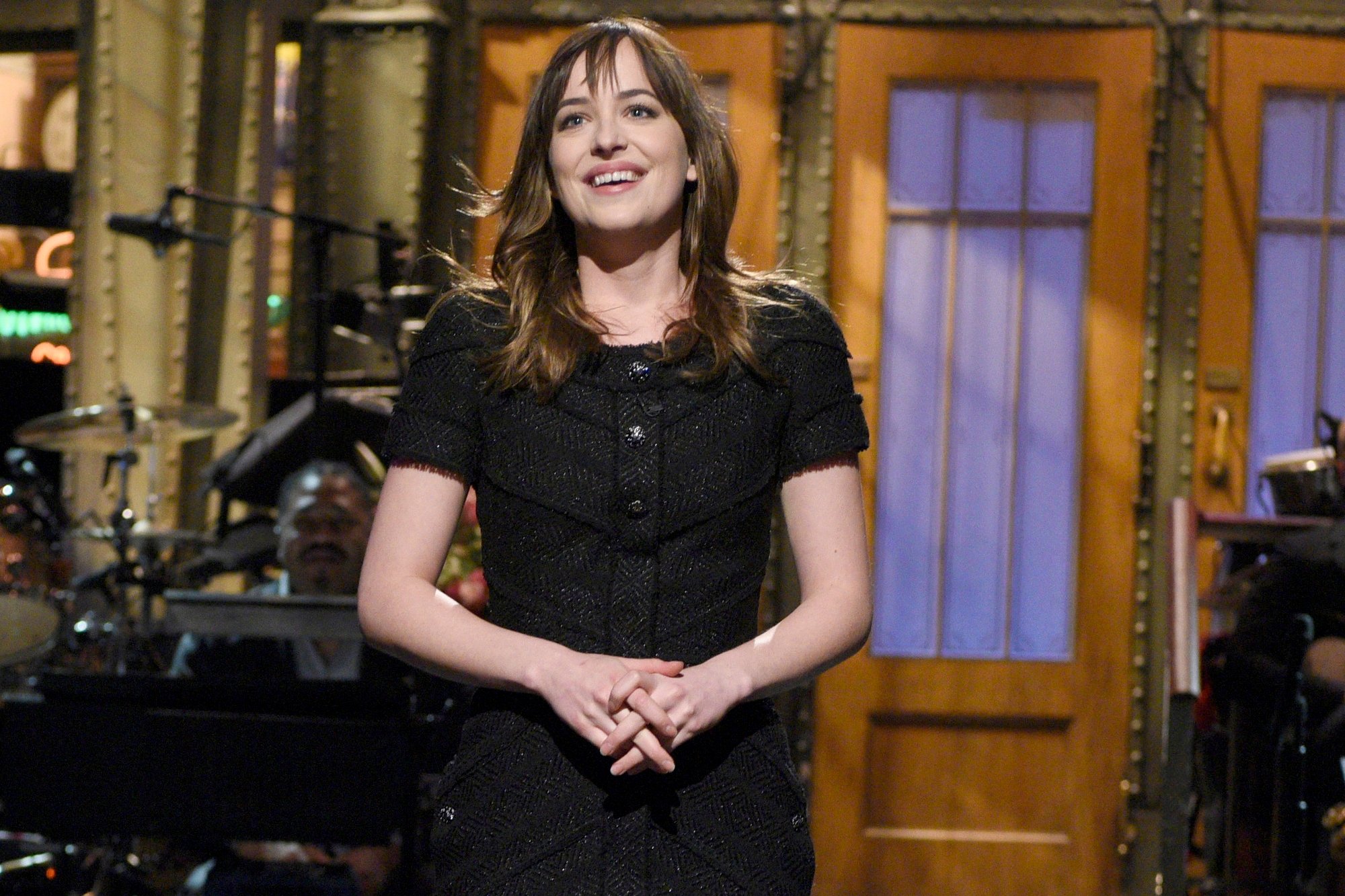 'Saturday Night Live' Dakota Johnson smiling and wearing a black dress on-stage. She's holding her hands together in front of her.