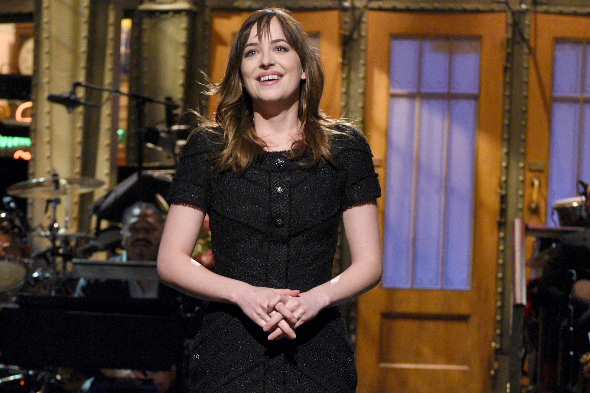 'Saturday Night Live' Dakota Johnson smiling and wearing a black dress on-stage. She's holding her hands together in front of her.