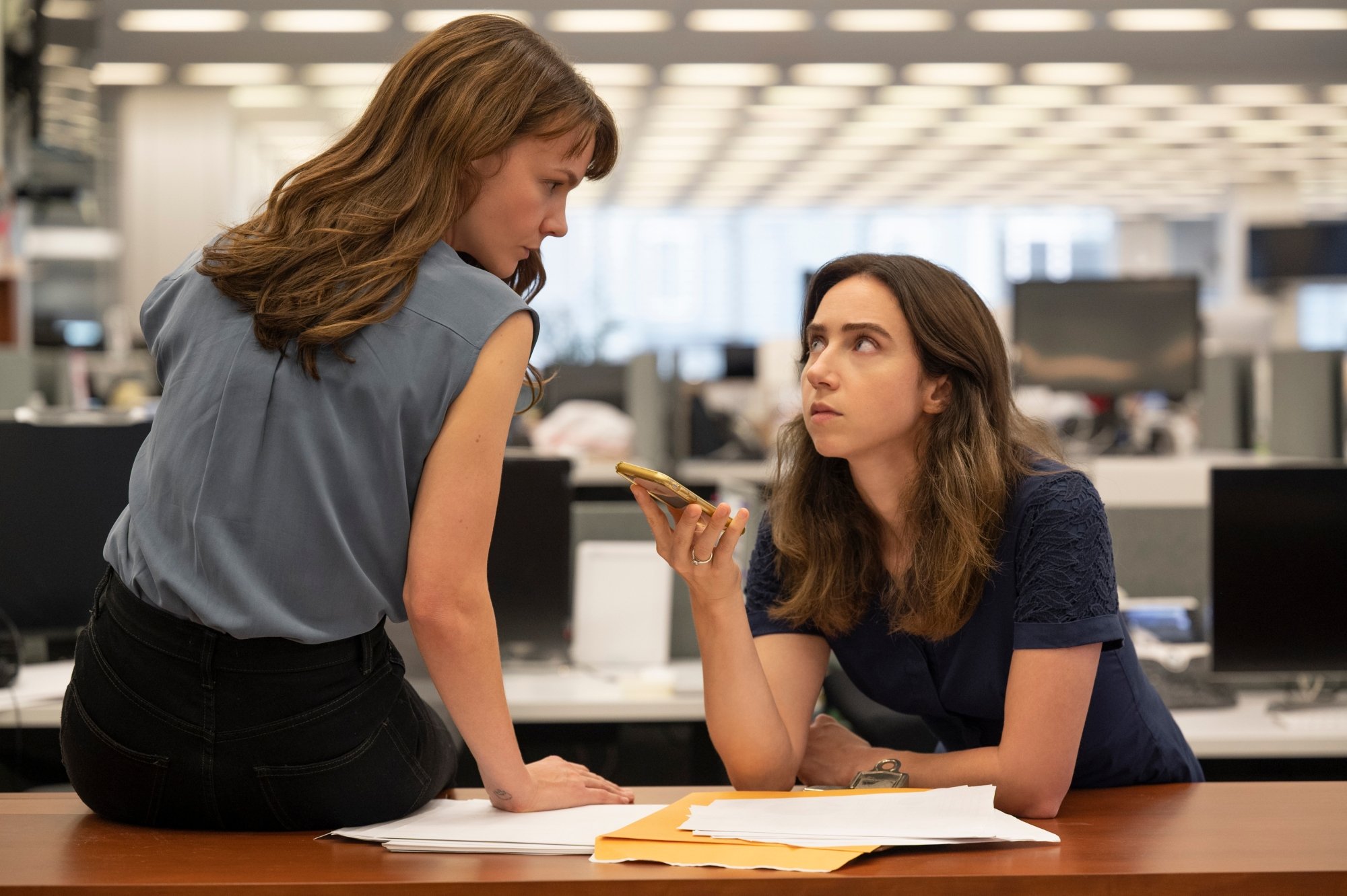 'She Said' Carey Mulligan as Megan Twohey and Zoe Kazan as Jodi Kantor. Kazan is leaning over a desk with paperwork on it. She's holding her phone while looking up at Mulligan, who is sitting on the desk, looking at her.