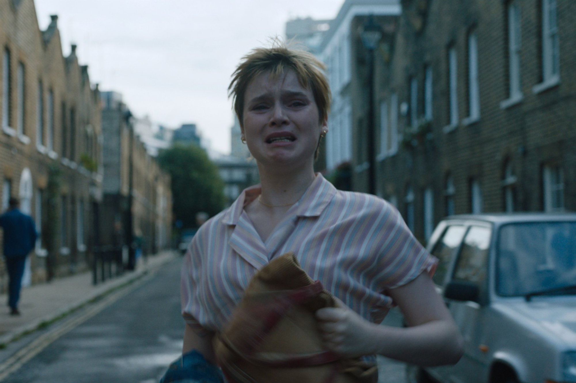 Lola Petticrew as Young Laura Madden in She Said, directed by Maria Schrader. running down the street holding a brown bag, while crying.