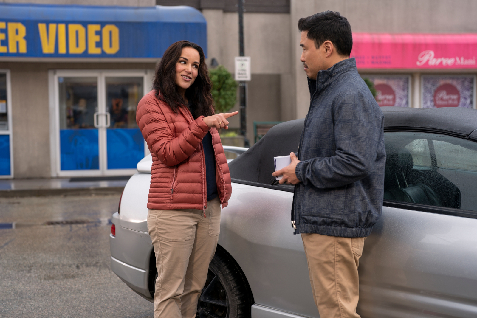 Melissa Fumero and Randall Park as Eliza and Timmy in 'Blockbuster' on Netflix for our article about shows like it. Eliza is pointing at Timmy in front of his car, and the store is in the background.