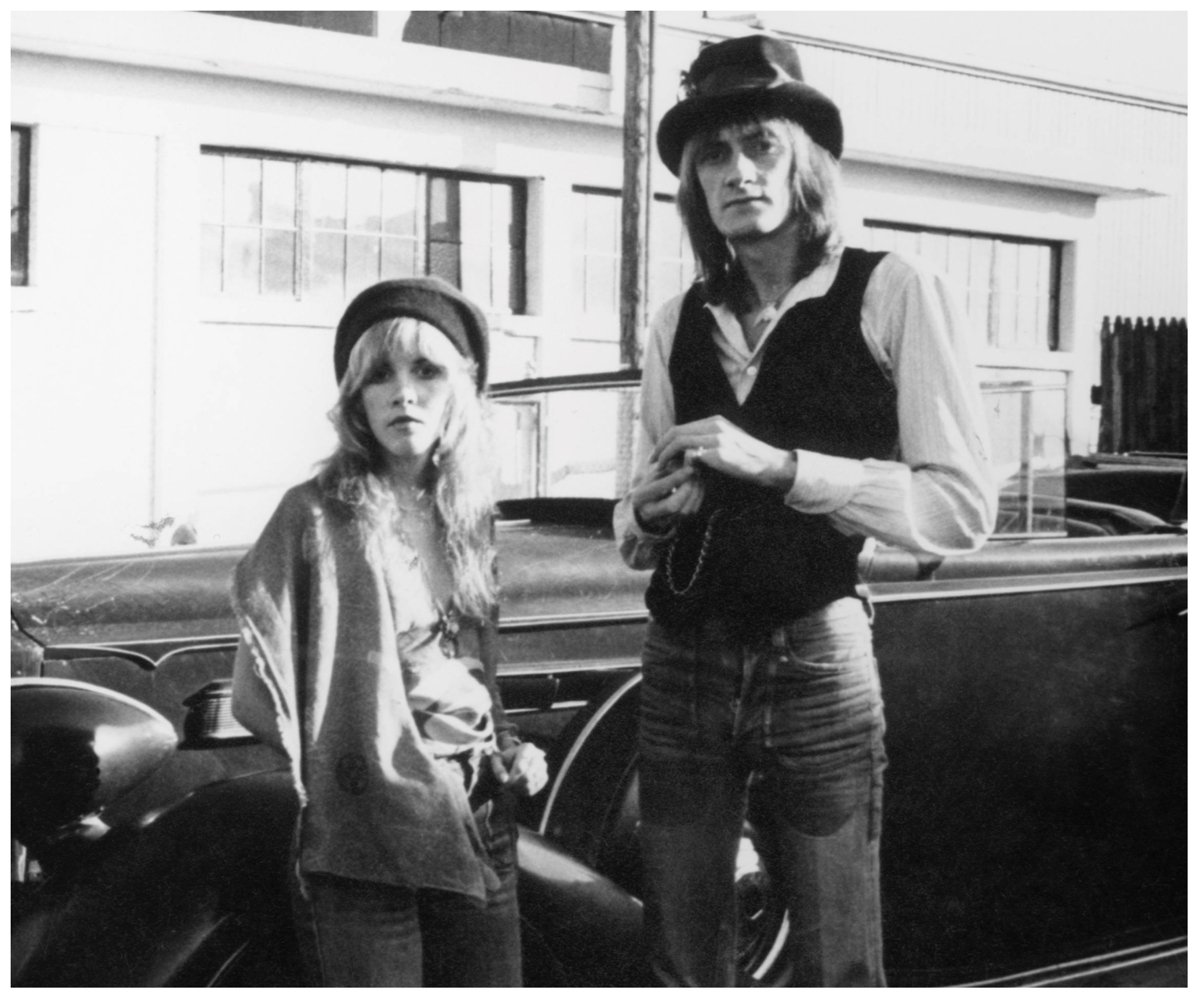 A black and white photo of Stevie Nicks and Mick Fleetwood, who had an affair while touring with Fleetwood Mac, posing together in front of a car.