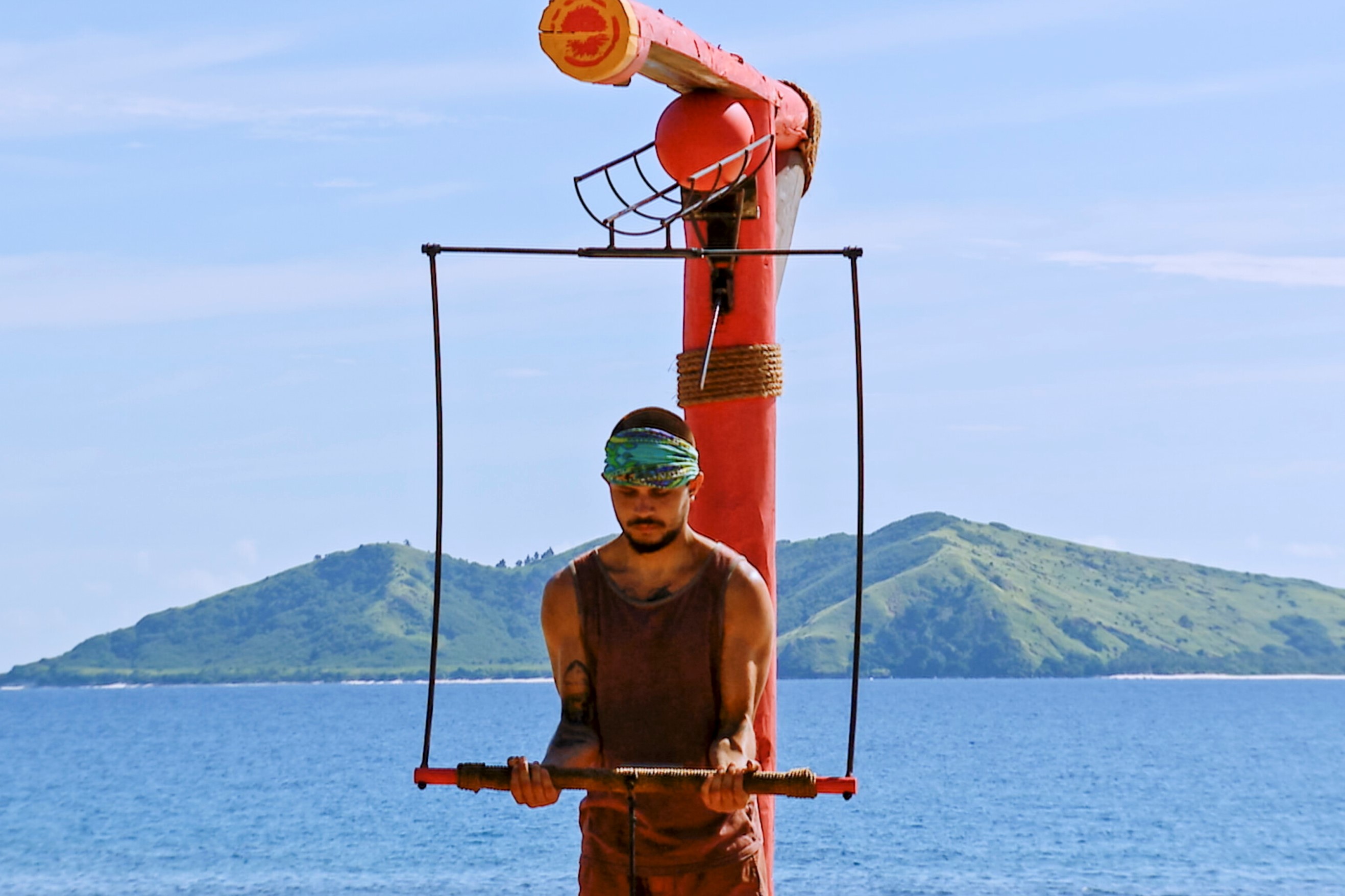 Jesse Lopez, who stars in 'Survivor 43' Episode 9 on CBS, competes in the Immunity Challenge, which includes holding a horizontal bar. Jesse wears a faded red tank top, red shorts, and the blue 'Survivor' merge buff.